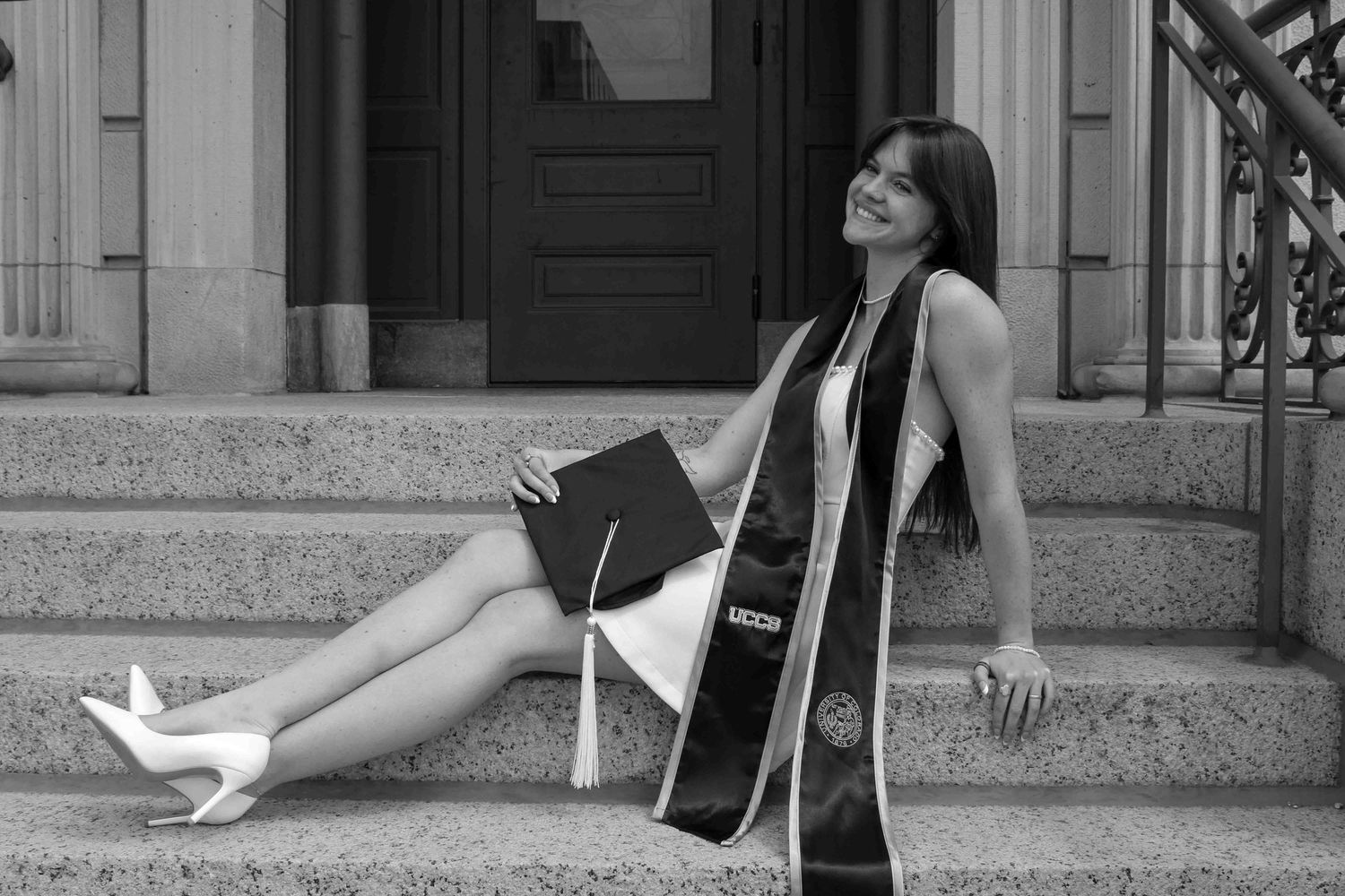 Black and white graduation photo of a young woman sitting on steps holding her cap and diploma, wearing a stole.