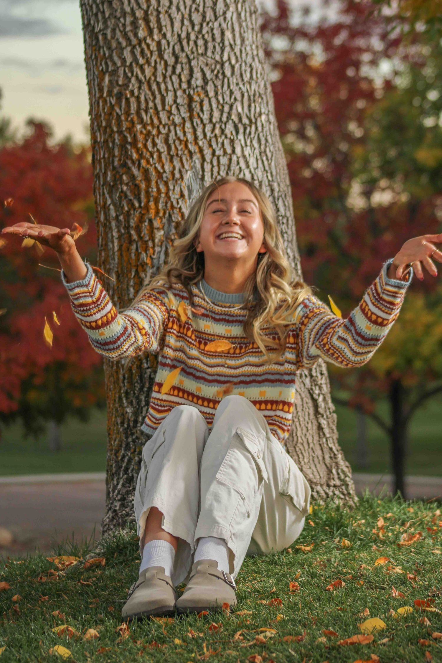 Someone sitting by a tree trunk on fallen autumn leaves wearing a patterned sweater with arms raised joyfully.