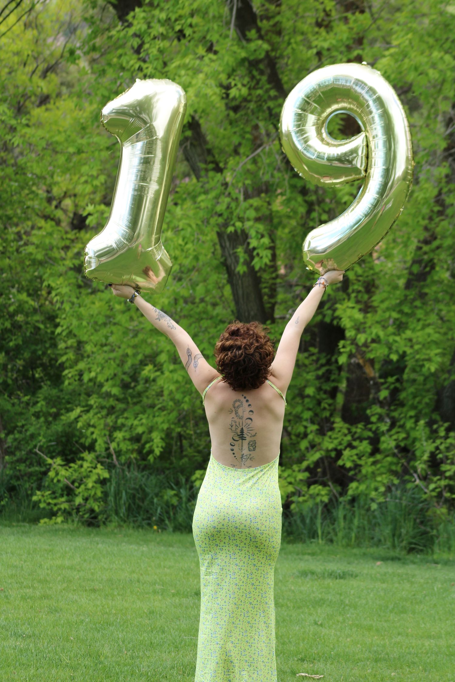 Person in green dress holding silver number 19 balloons overhead in a lush garden setting.