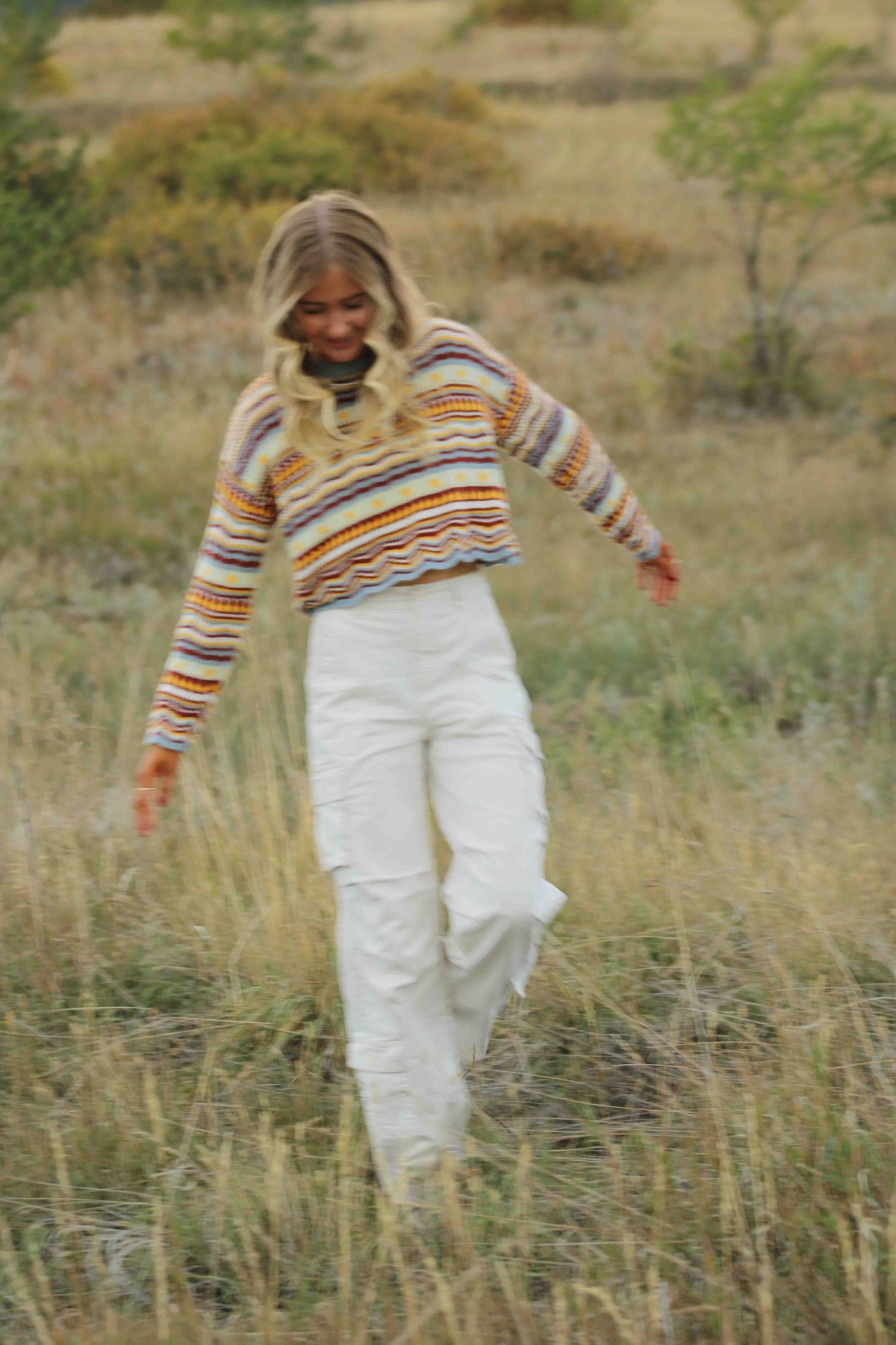 Person in striped sweater and white pants walking through tall grass in a field.
