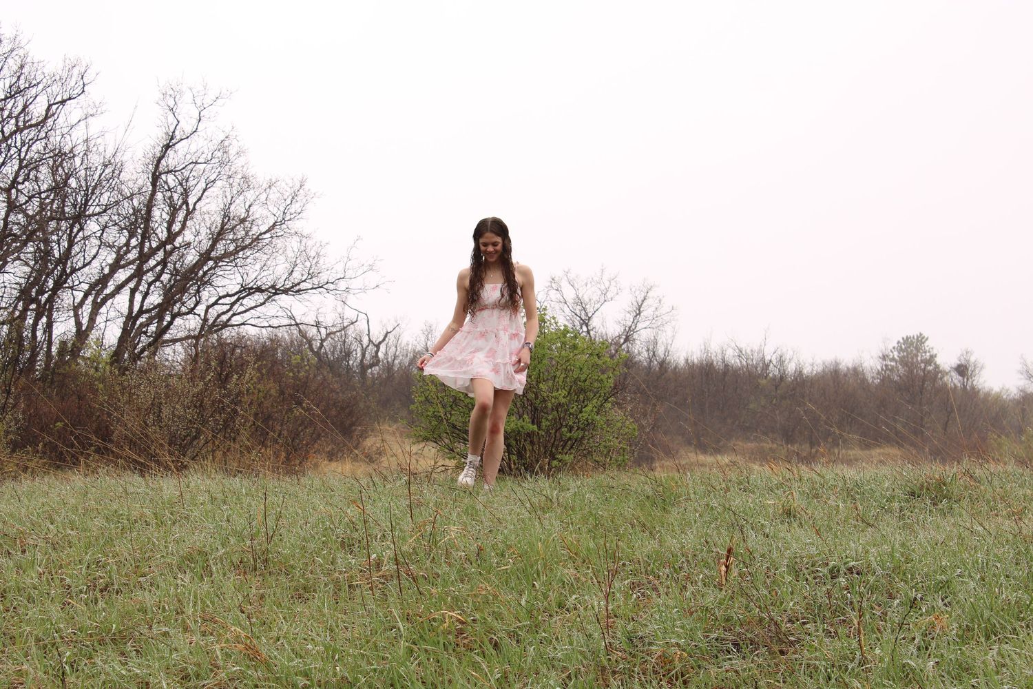 A person in a white dress stands in a grassy meadow with bare trees in the background.