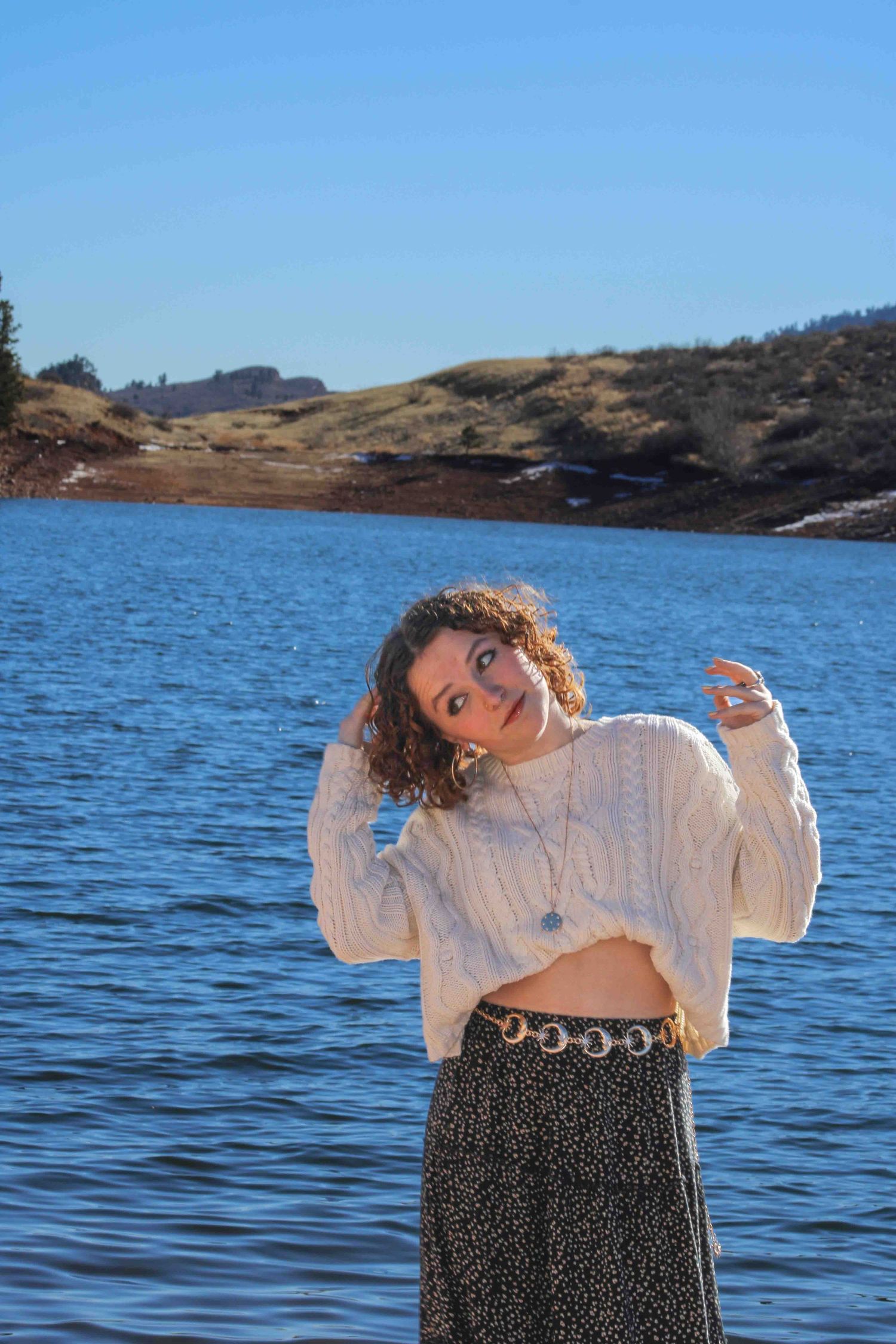 Dancing figure in white sweater and patterned skirt moves playfully against blue lake backdrop.