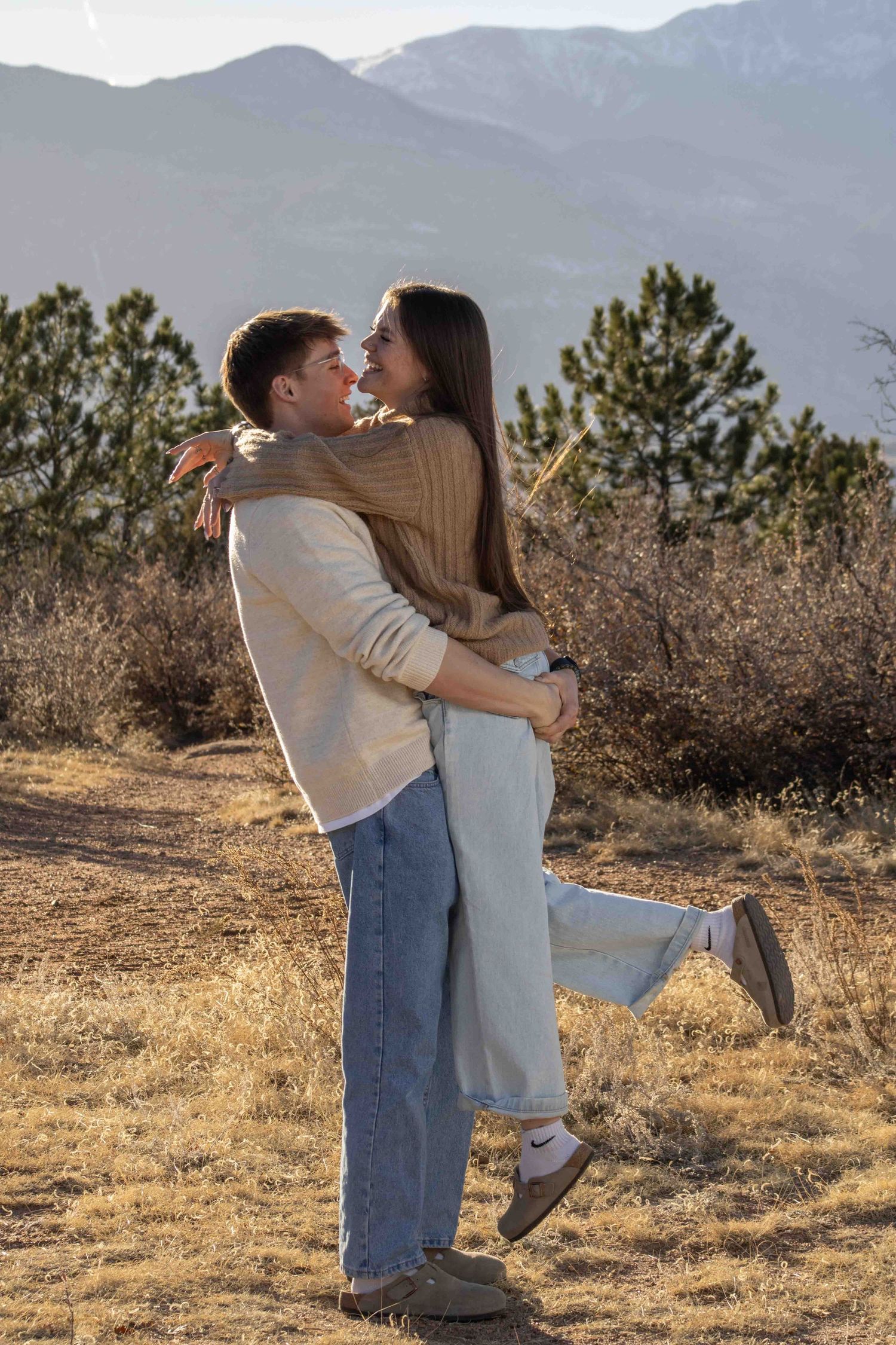 A playful couple embraces against mountain backdrop with one person lifted off the ground.