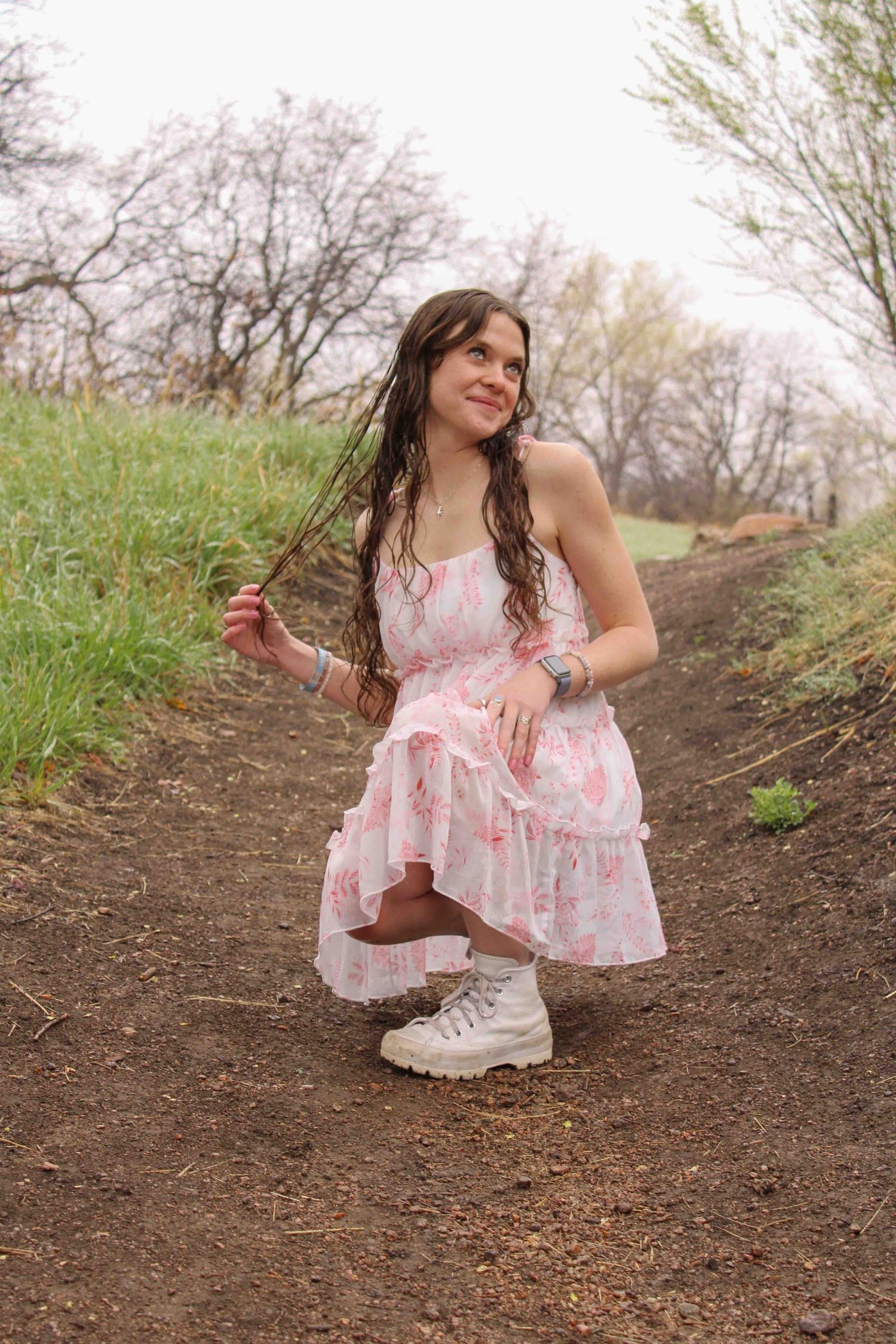 Someone wearing a floral pink sundress and white sneakers poses crouched on a dirt path surrounded by spring greenery.