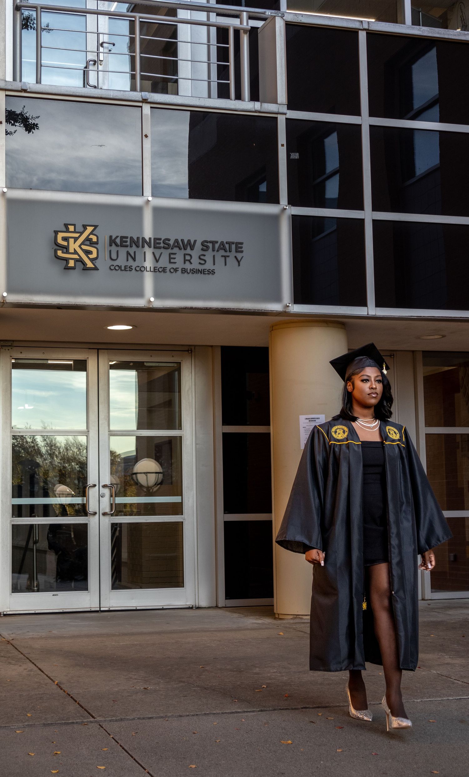 Graduate in traditional academic regalia standing in front of Kennesaw State University entrance.