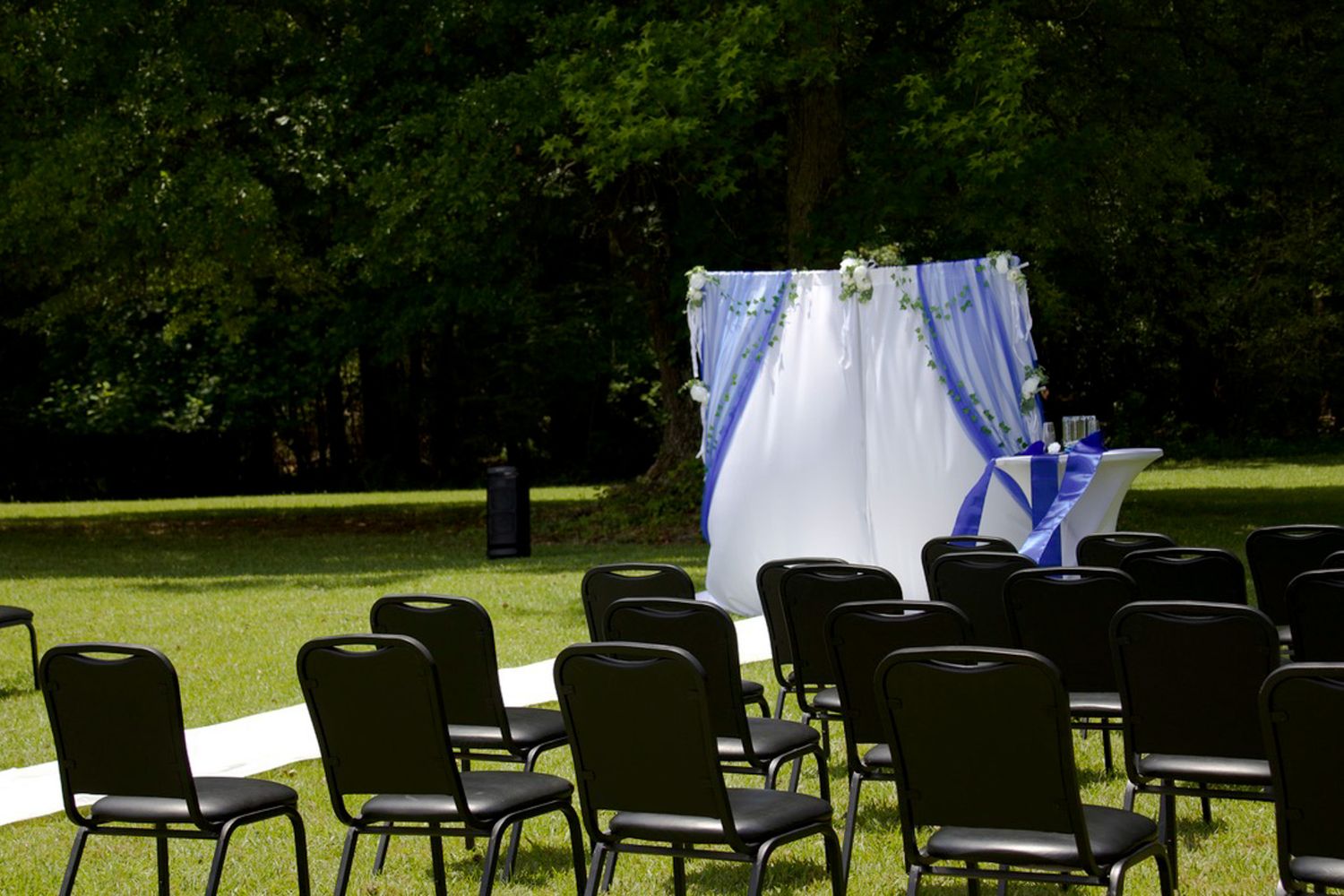 Outdoor wedding ceremony setup with black chairs and white draped arch with blue accents in a green garden setting.