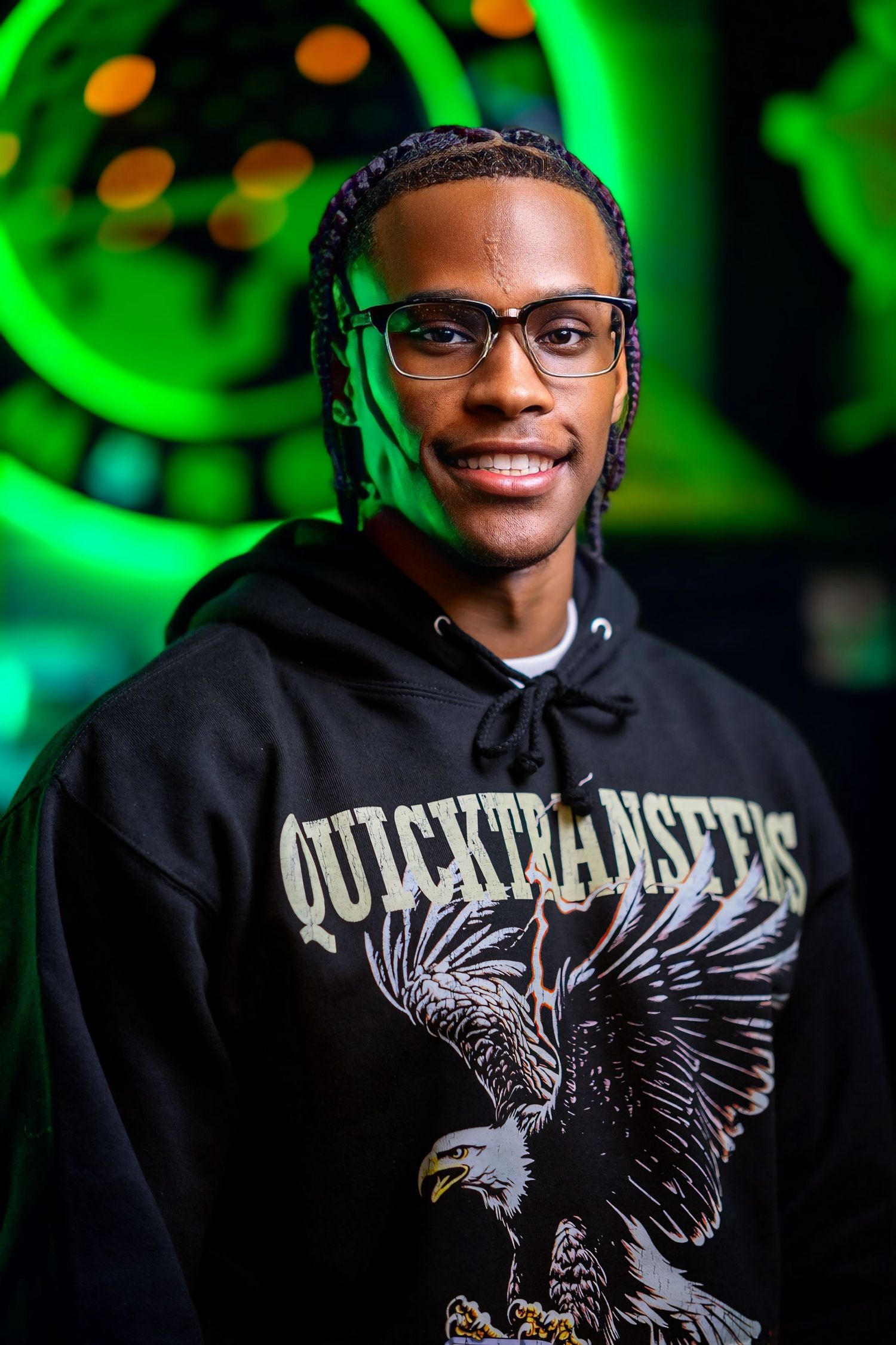 Fashionable young person in glasses wearing a black OutKast Masters hoodie with eagle graphic against green lights.