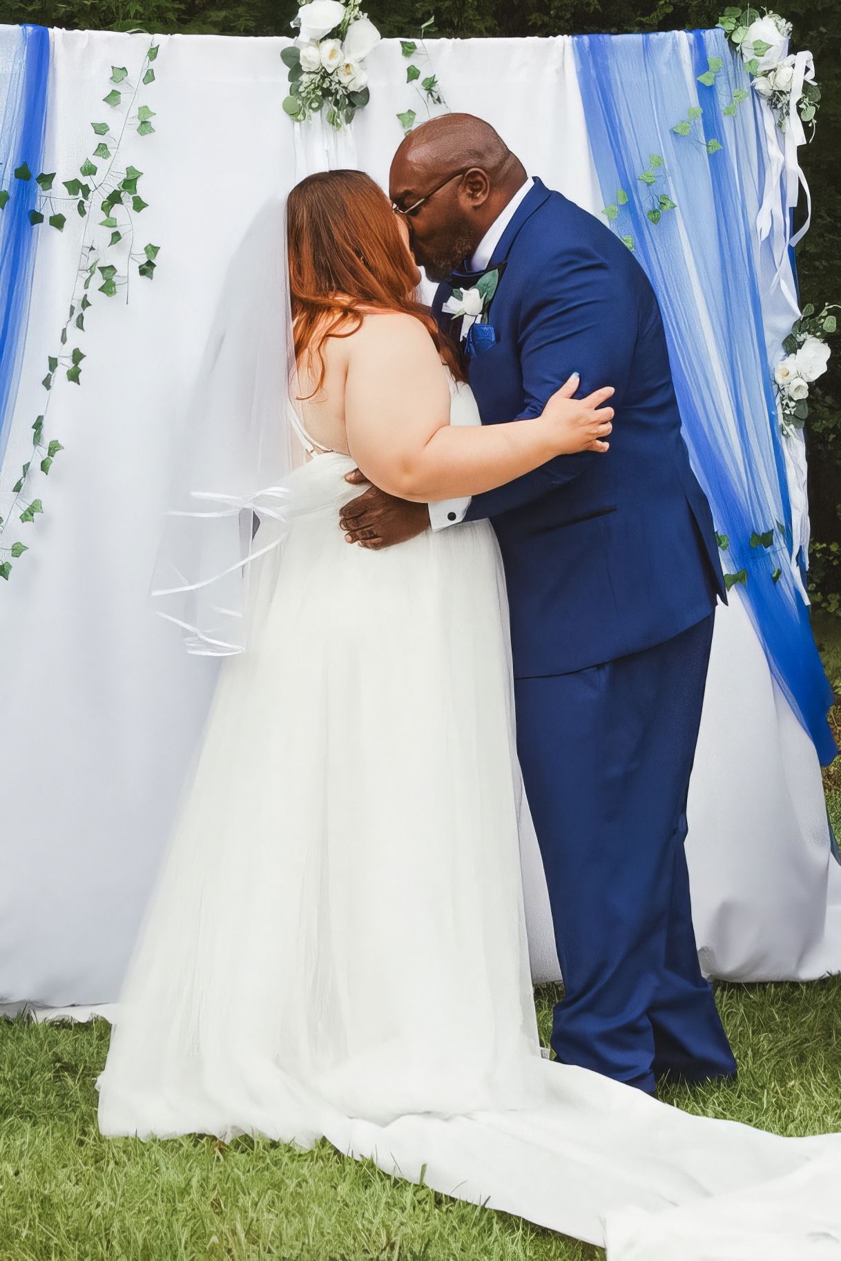 A couple shares their first kiss during an outdoor wedding ceremony under a white and blue draped arch decorated with greenery.