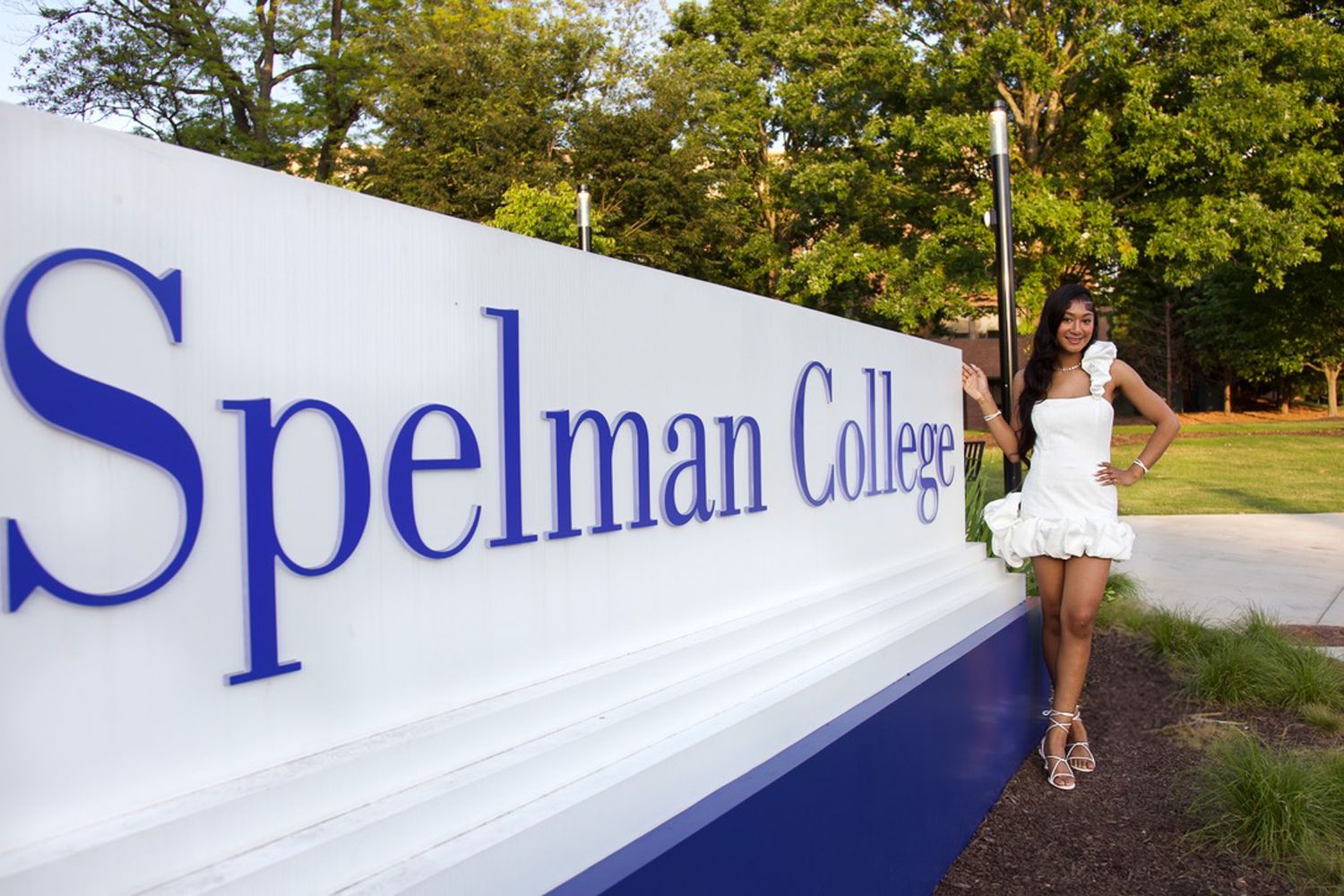 Student in a white dress stands next to Spelman College entrance sign on a sunny day.