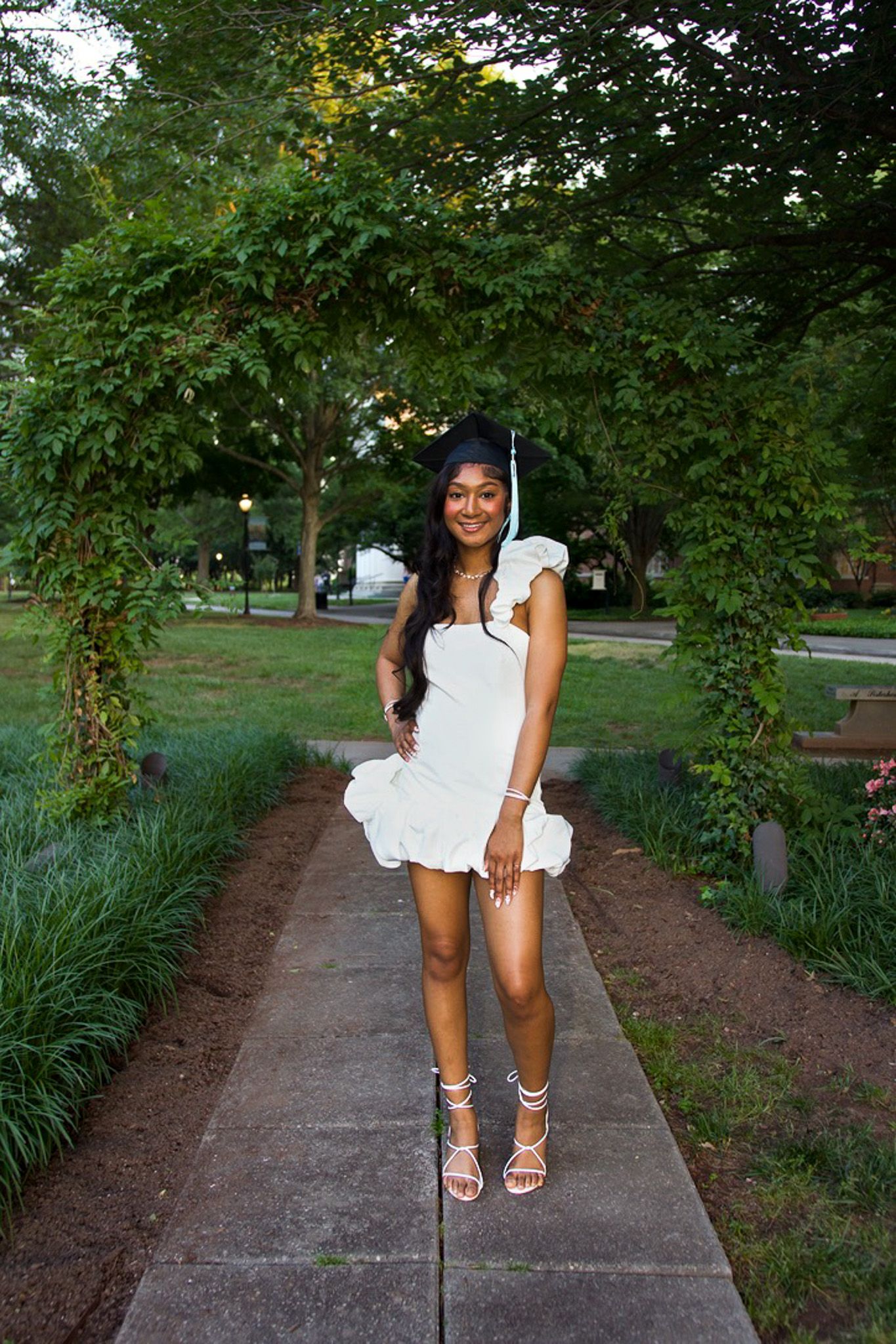 Woman in a white summer dress poses on a garden path surrounded by lush green trees and landscaping.