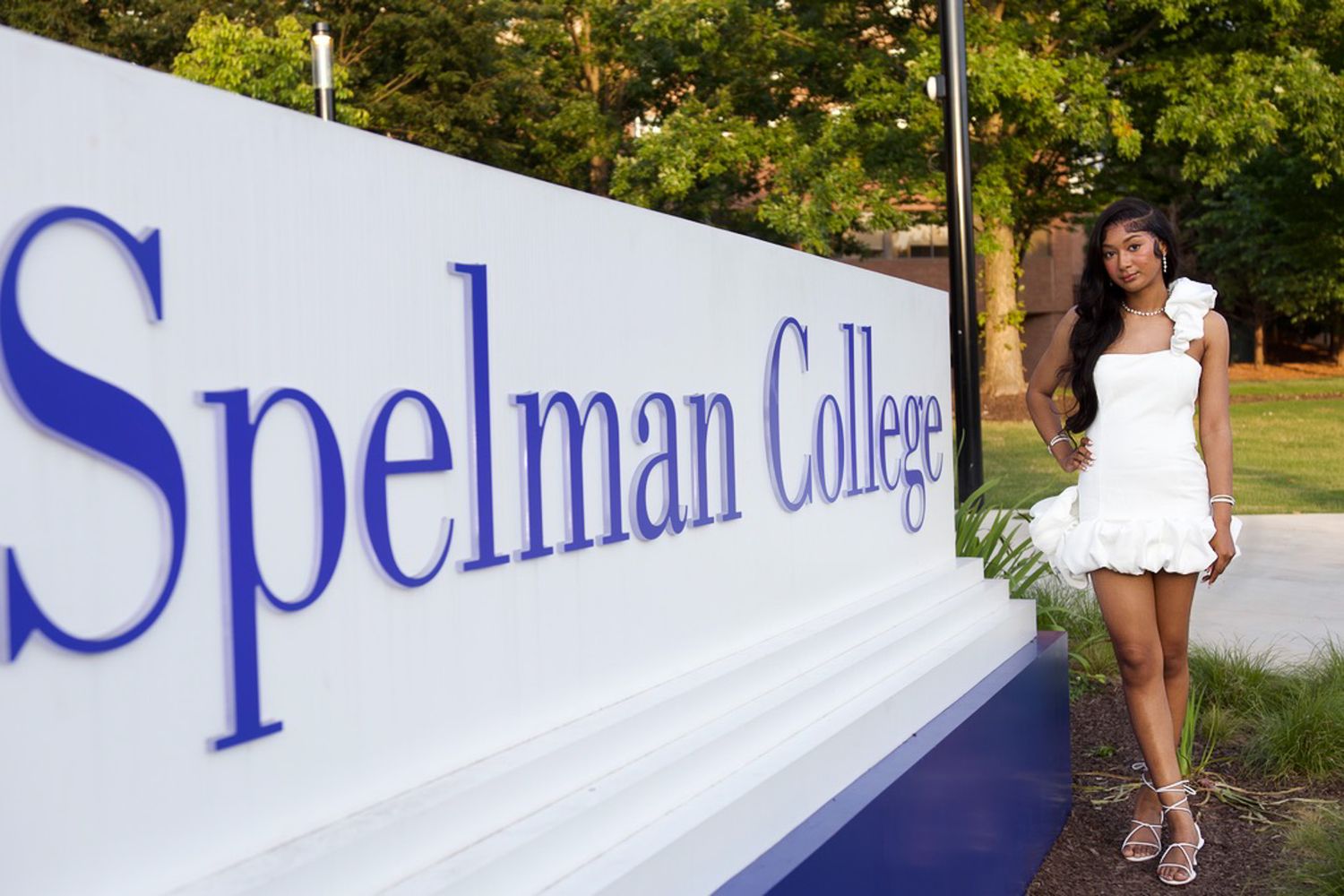 A student poses in a white dress next to the Spelman College entrance sign on a sunny day.