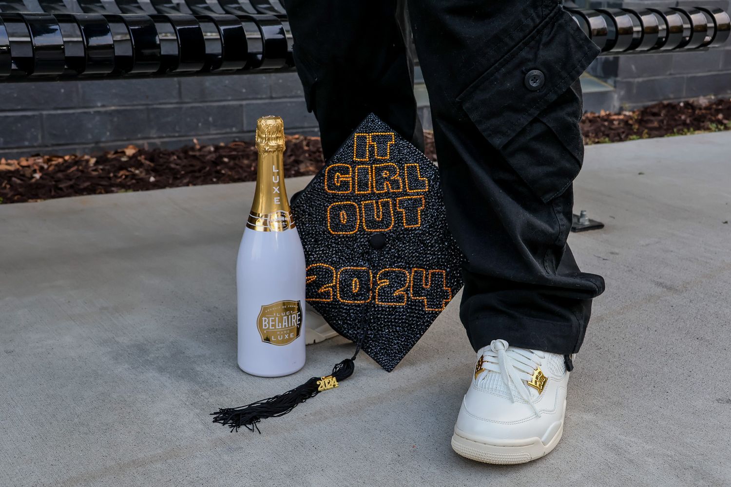 A graduation cap, white Nike sneaker, and celebratory champagne bottle arranged on concrete ground.