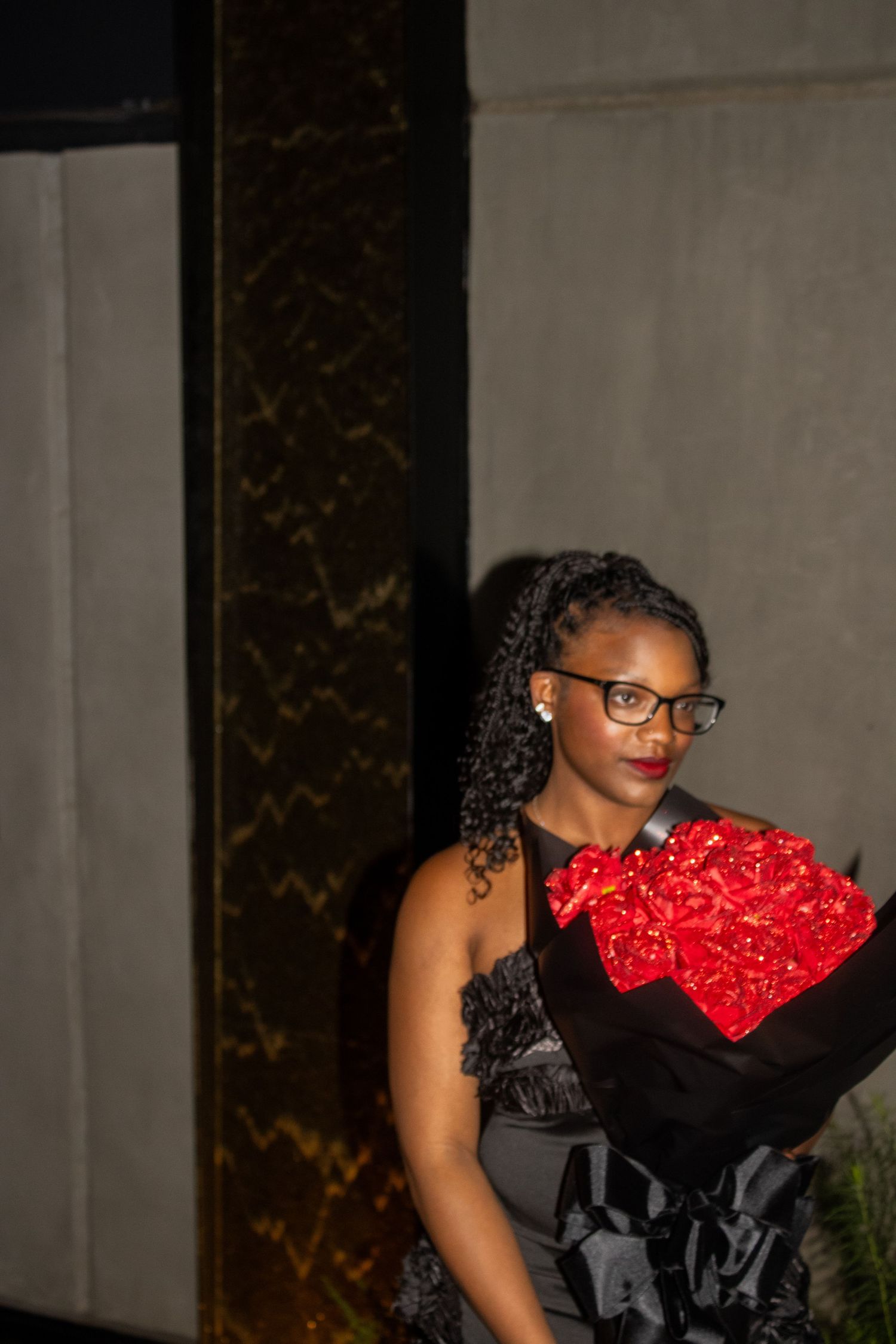 Woman in black dress poses with red rose bouquet against marble column backdrop.