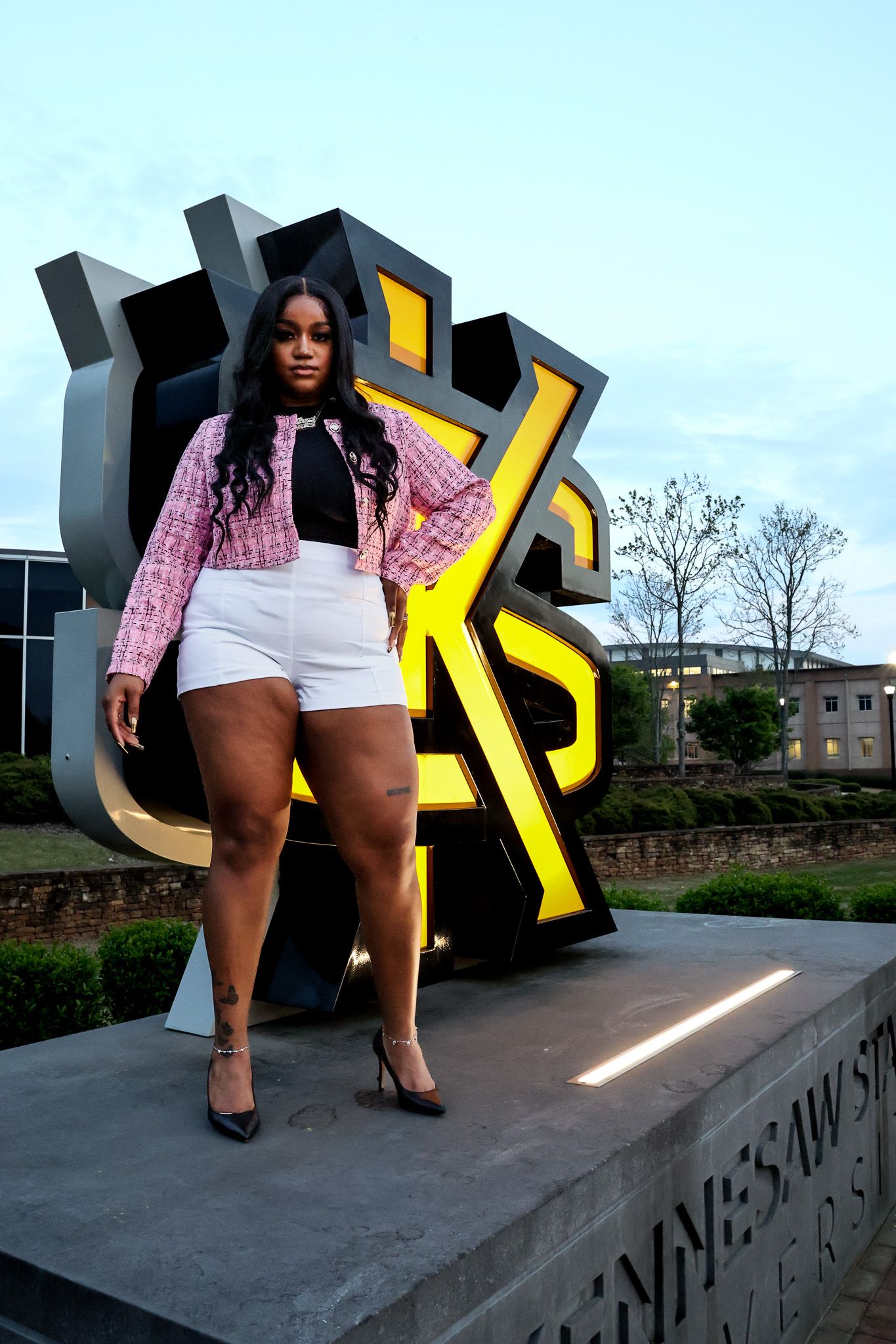 Pink tweed jacket paired with white shorts against large yellow university signage.