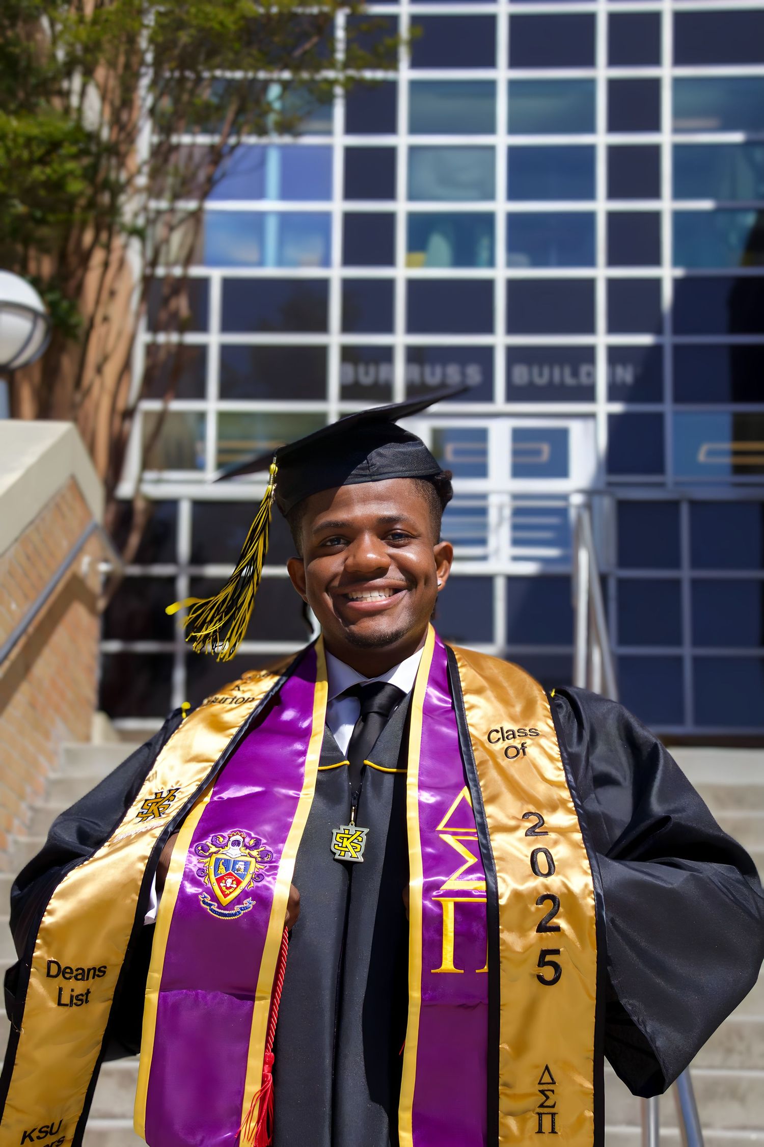 Graduate in cap and gown wearing purple and gold stoles smiles in front of campus building.