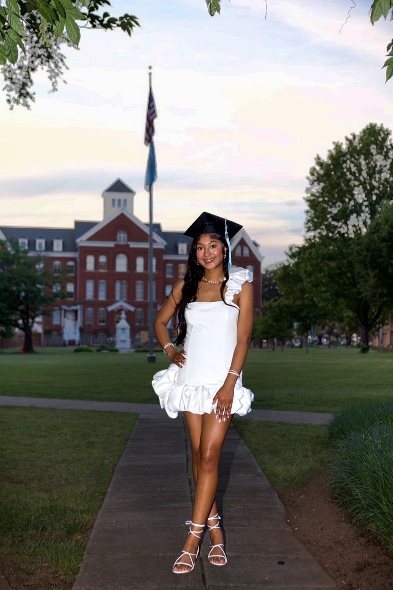 A white graduation cap and dress contrast against a traditional brick building with a flagpole at sunset.