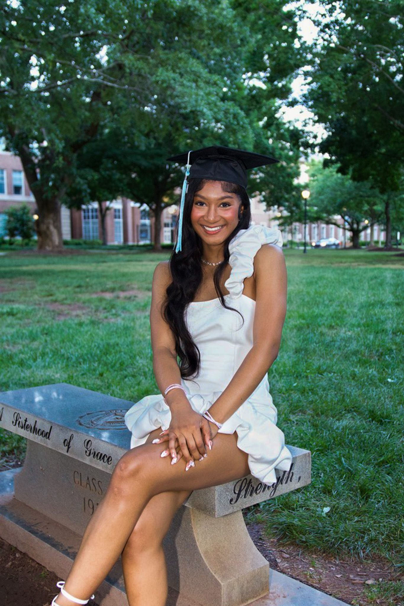 Graduate in white dress sitting on stone bench among green trees on campus grounds during graduation photoshoot.