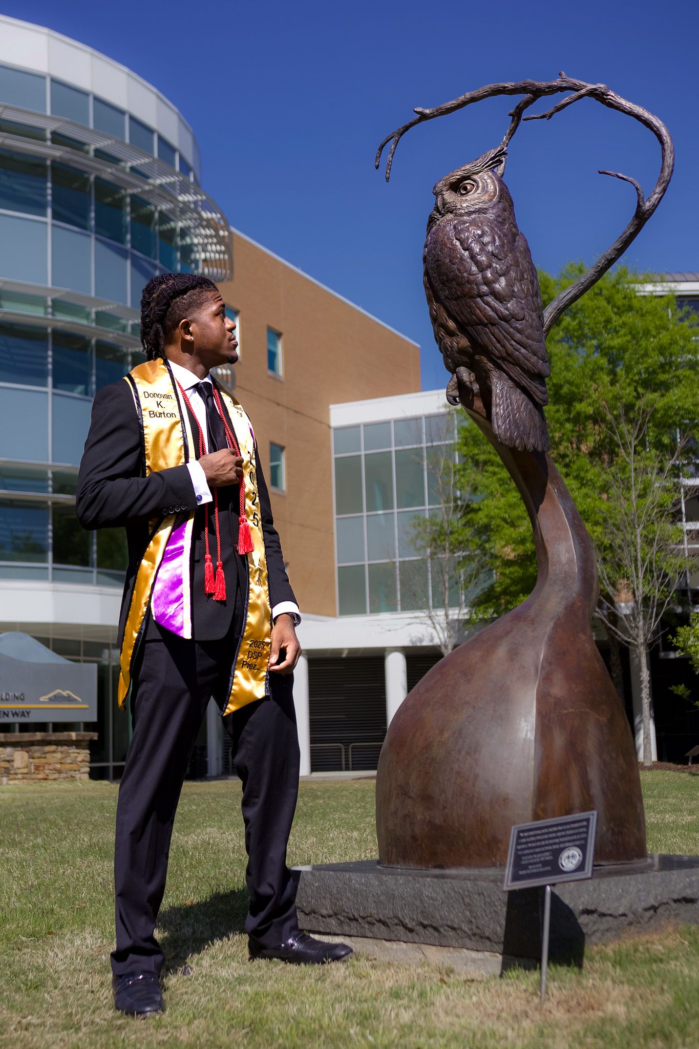 Graduate in academic regalia poses next to large bronze owl sculpture on campus.