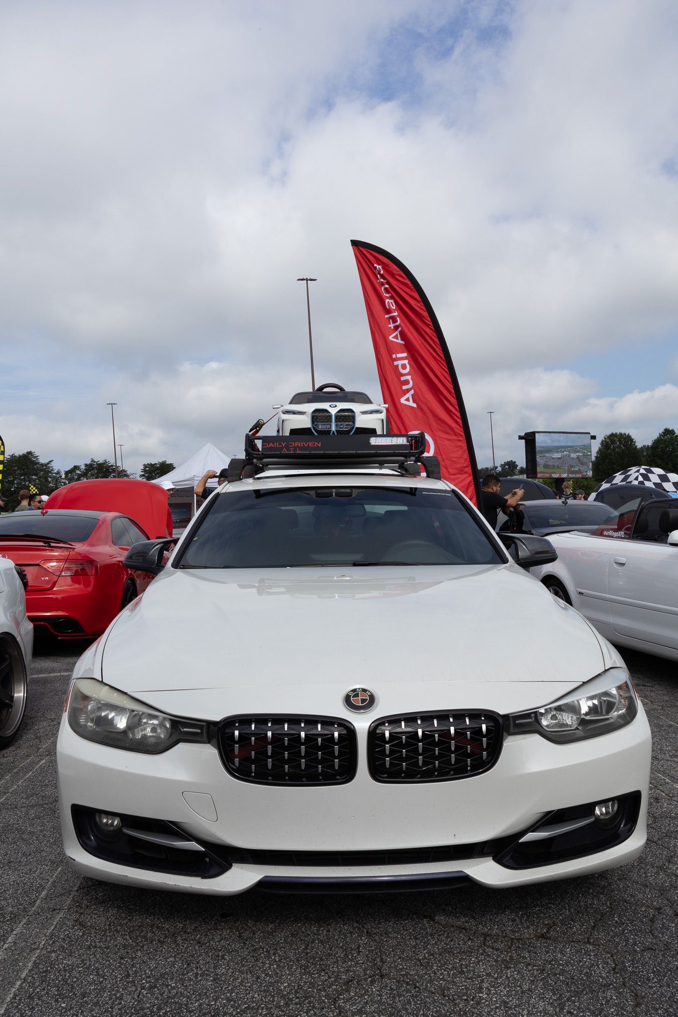 Front view of a white BMW 3 series with roof rack and red banner flag in a parking lot.