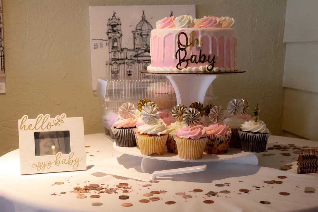 Pink baby shower cake and cupcakes displayed on white tiered stand with decorative elements.