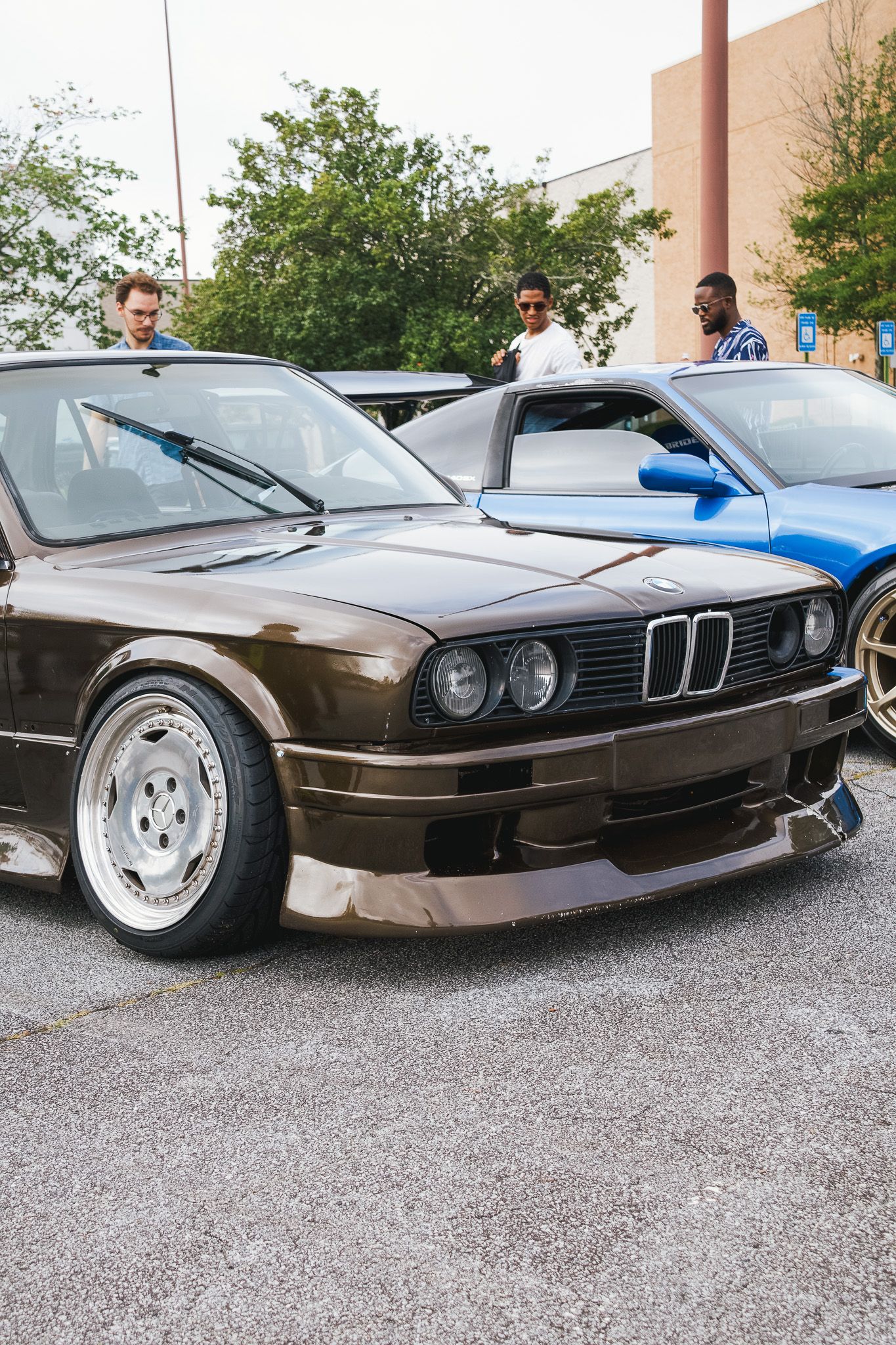 Modified brown BMW E30 with custom wheels and body kit in a parking lot.