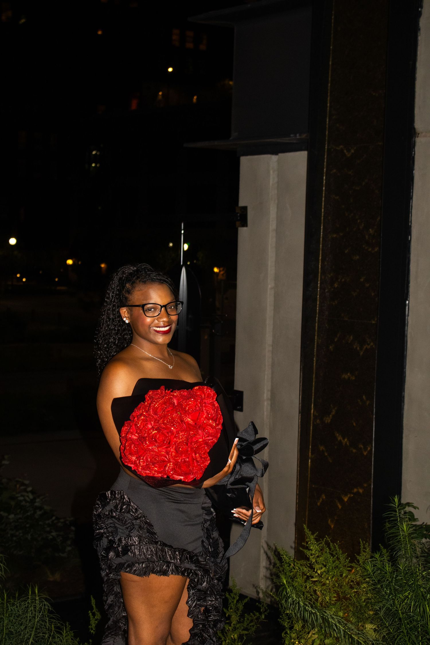 Elegant woman in black dress holding vibrant red roses bouquet at night against white wall backdrop.