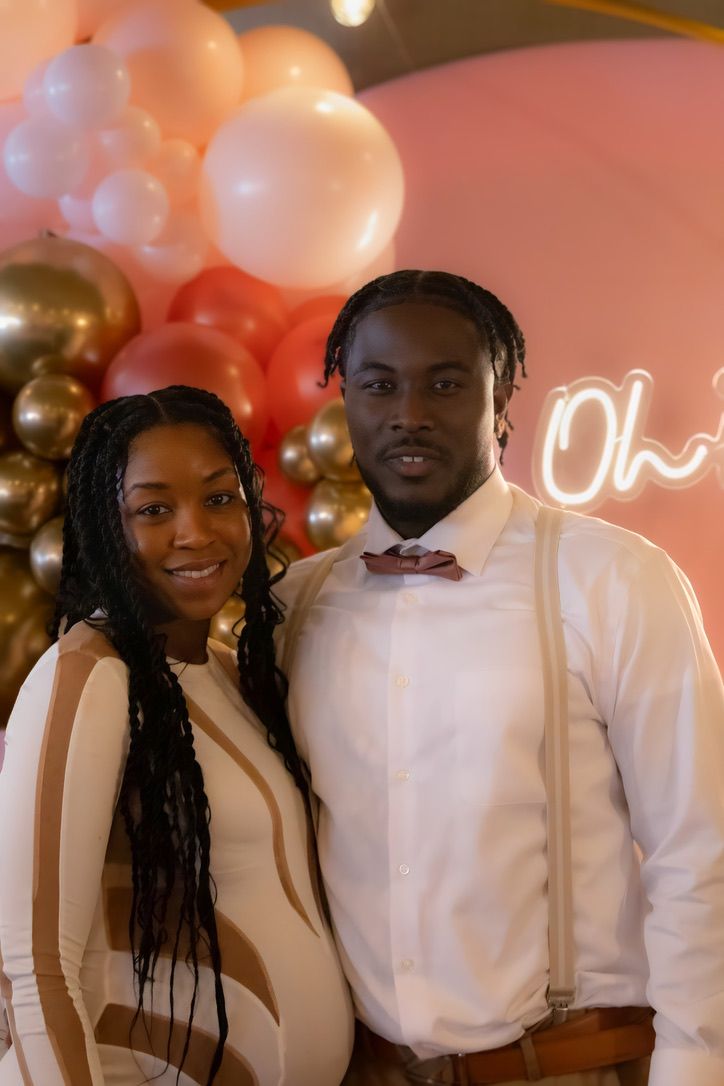 Expecting couple poses in formal attire against balloon arch with neon sign.