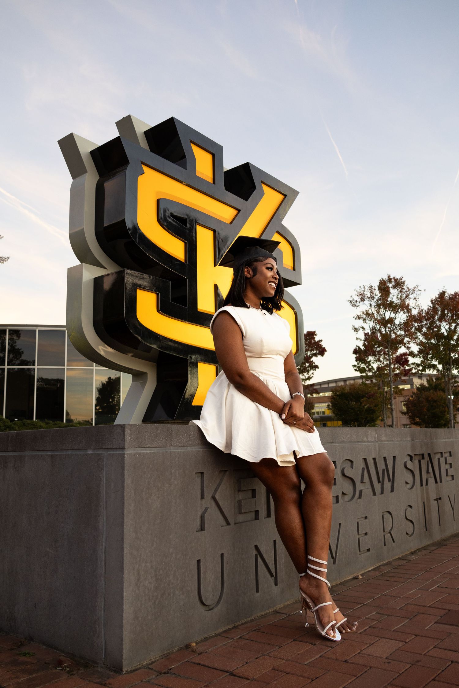 White-dressed graduate posing by large yellow KSU logo sculpture at Kennesaw State University.