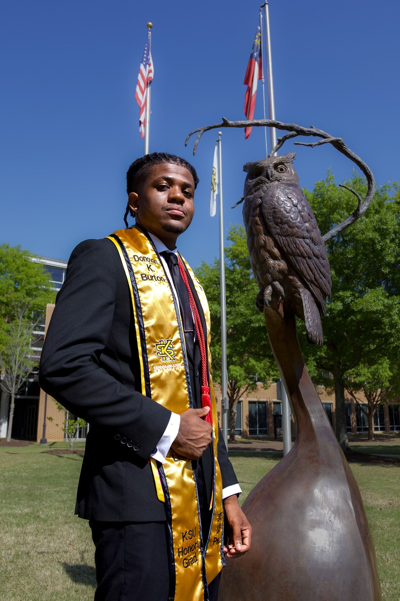 Graduate in academic regalia stands by owl statue with American and state flags.