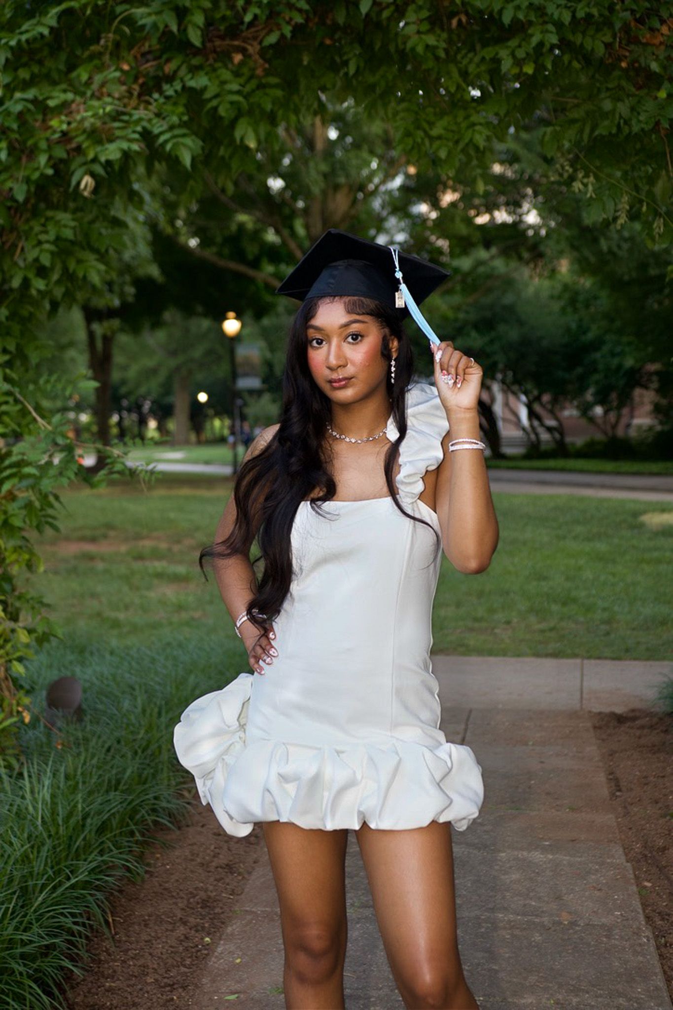 Graduate in white mini dress with ruffled hem poses on walkway surrounded by lush greenery on sunny day.