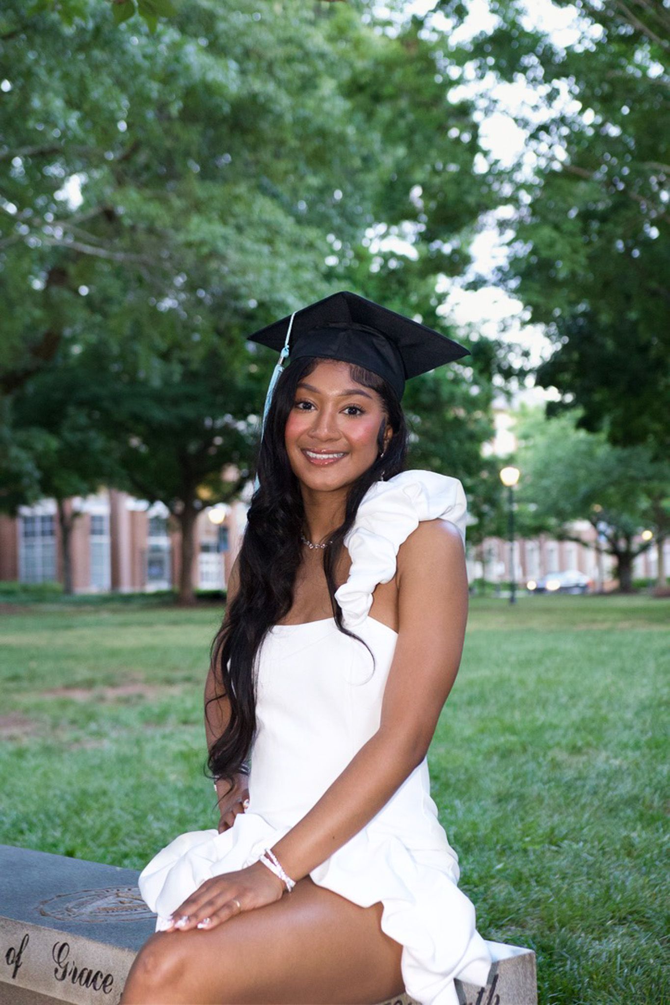 Graduate in white dress and cap sits on stone structure outdoors with green trees in background.