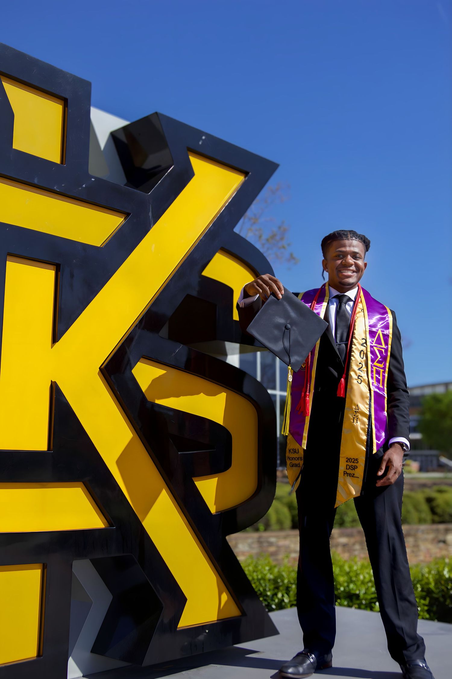 Graduate in academic regalia poses triumphantly next to large yellow university logo sign.