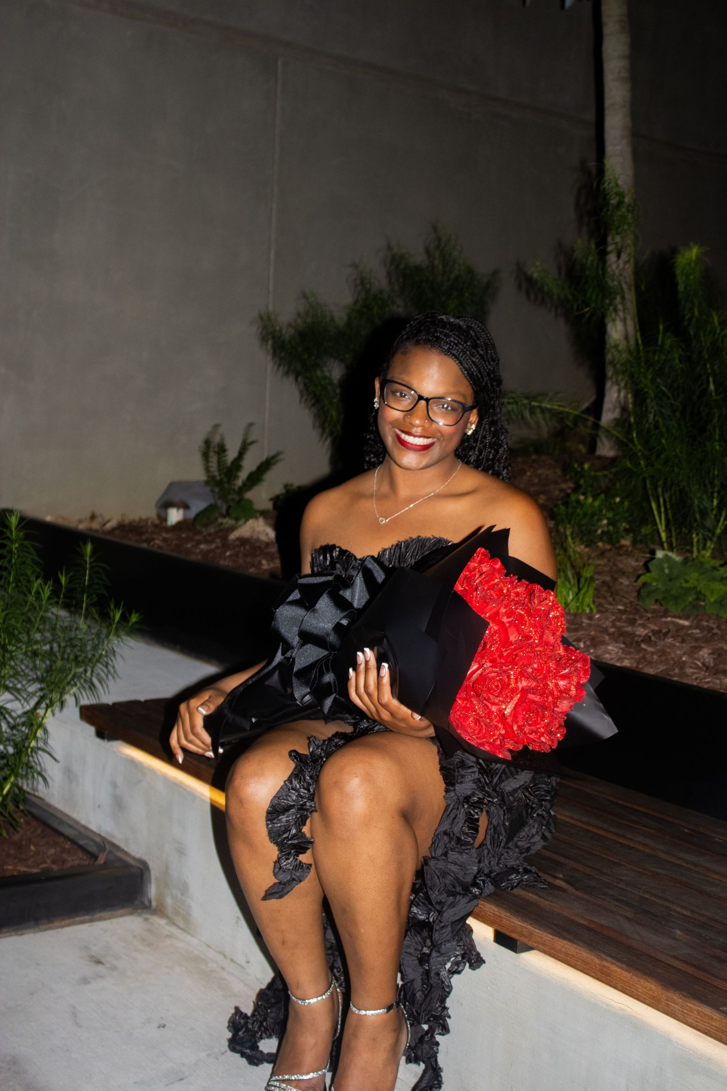 Woman in black dress sits on outdoor bench while holding red rose bouquet.