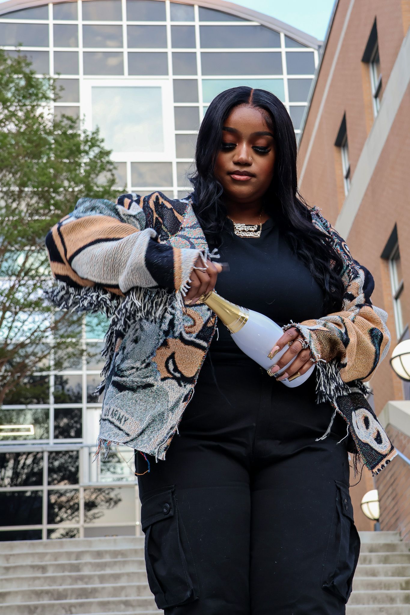 Stylish streetwear outfit with graphic print jacket and champagne bottle against architectural backdrop.