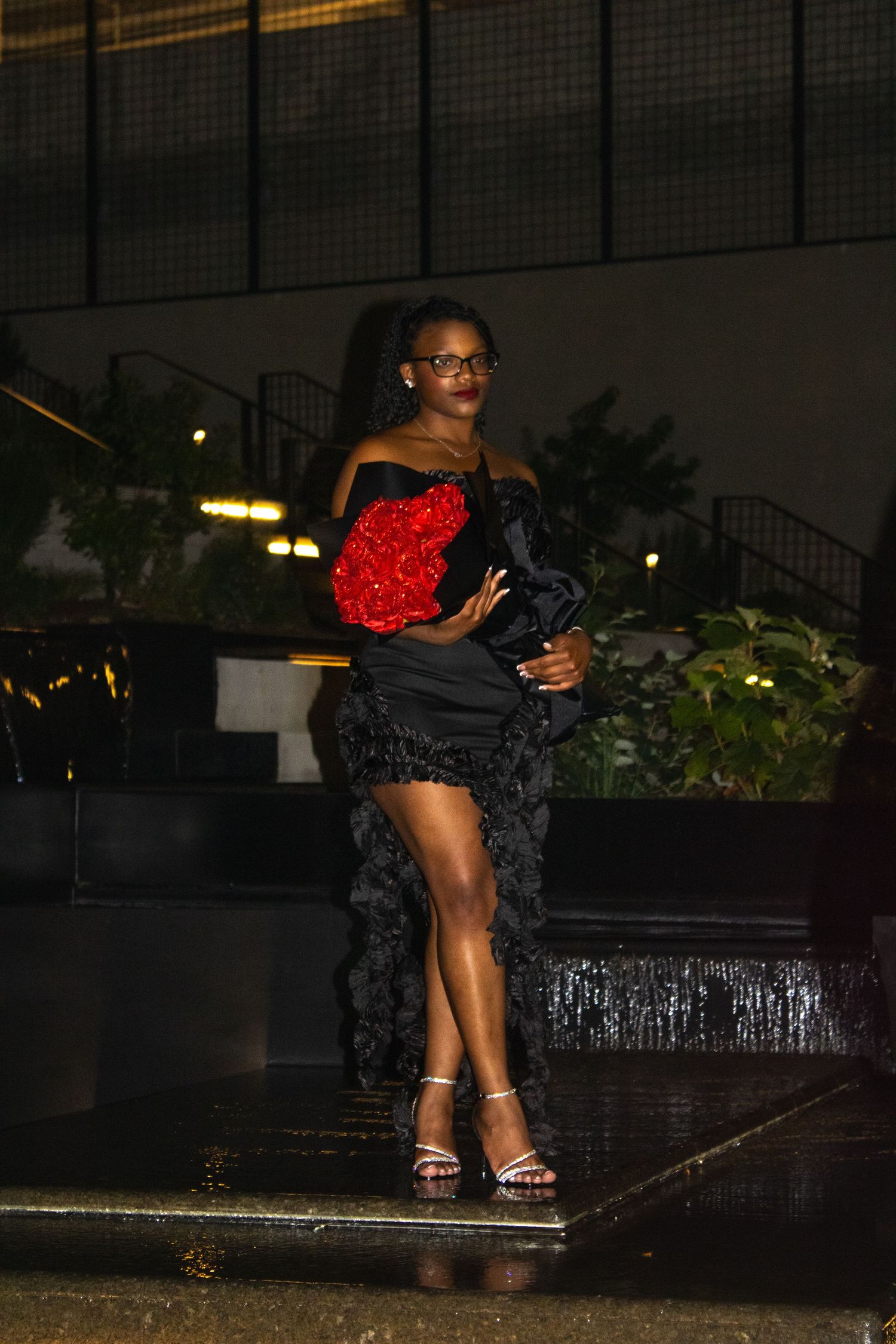 Woman poses in black dress and heels while holding red rose bouquet outdoors.