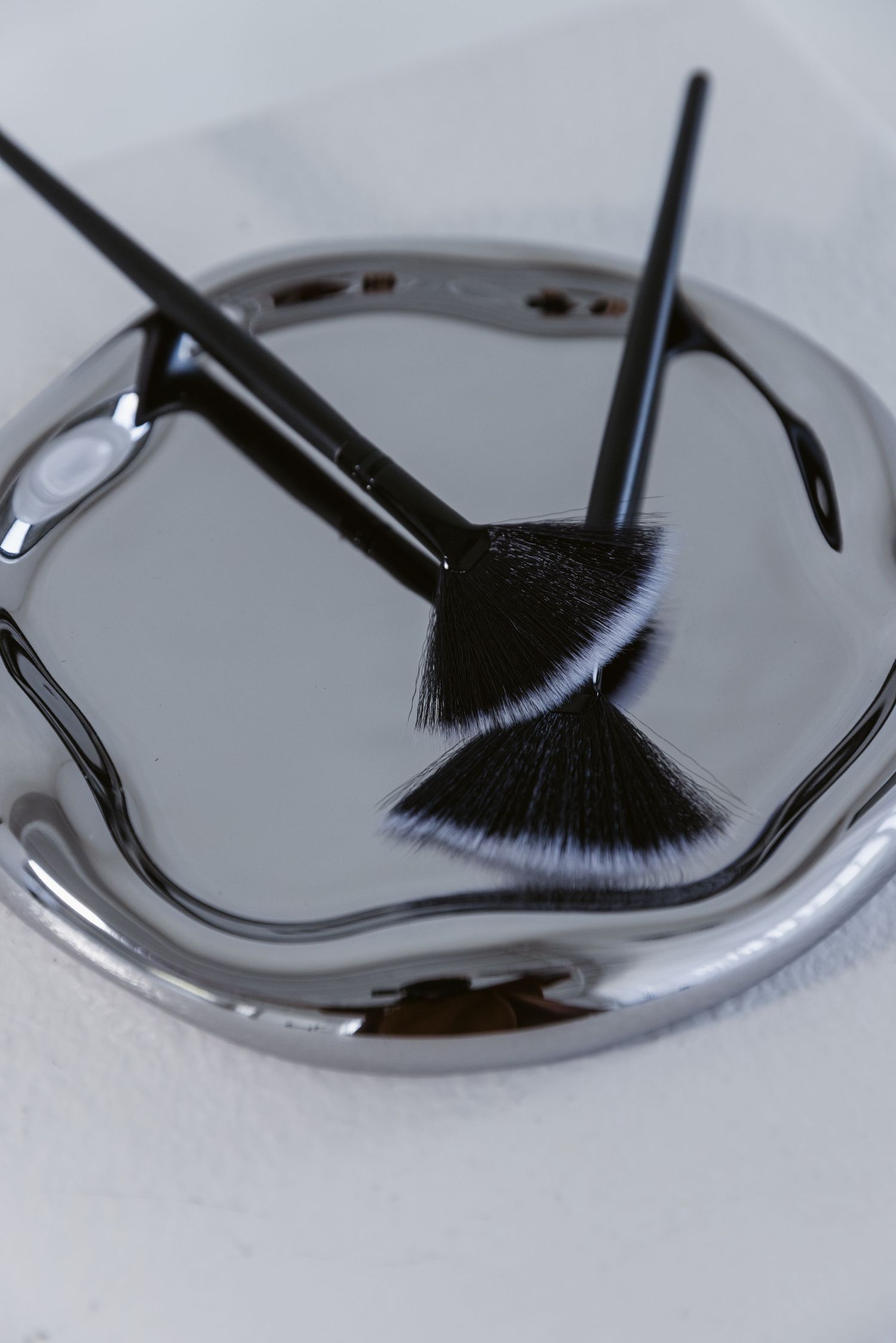 Two black fan makeup brushes soaking in water inside a silver metallic sink drain.