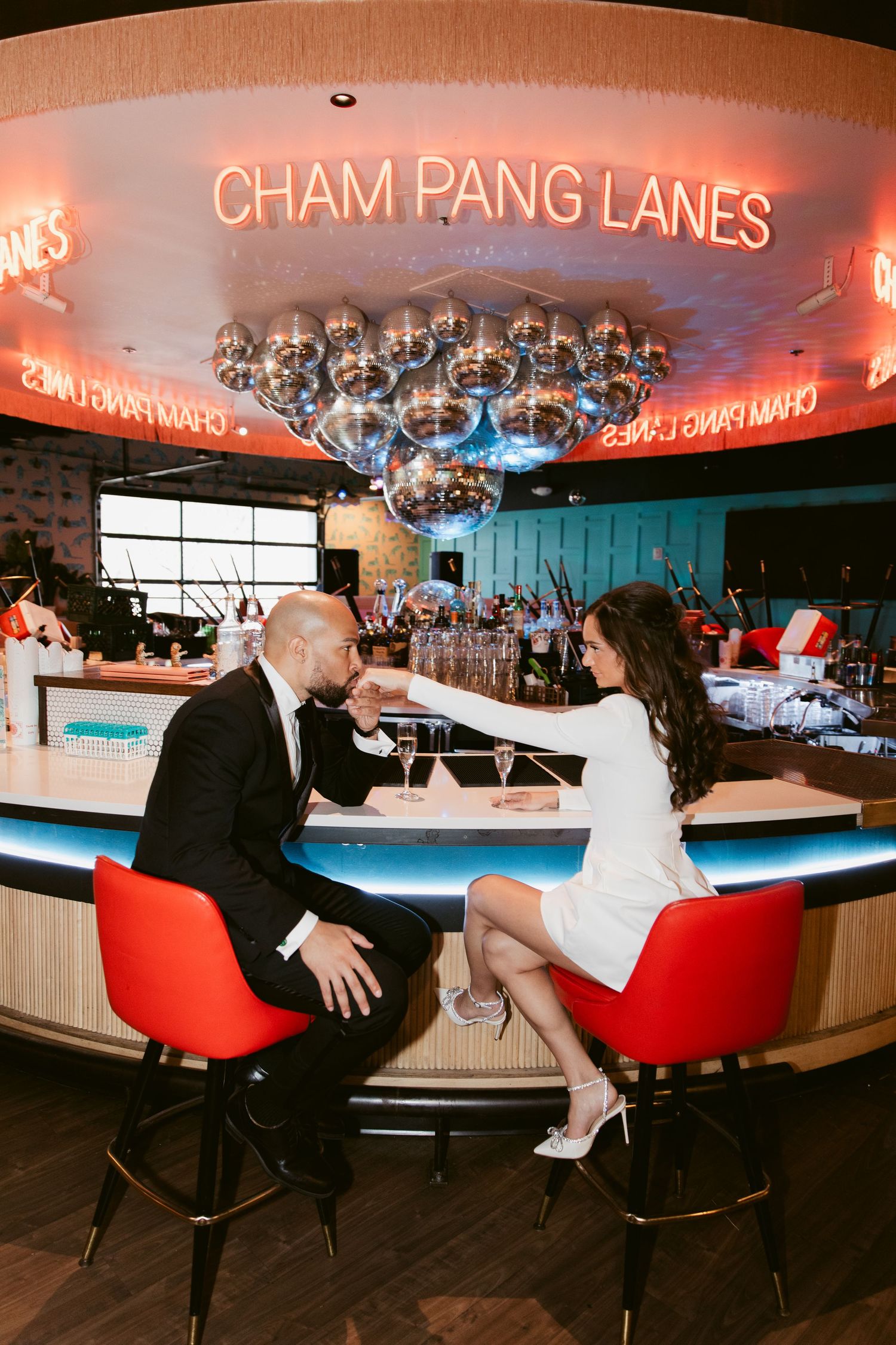 A couple sits at a retro bar under neon 'Cham Pang Lanes' signage with disco ball lighting.