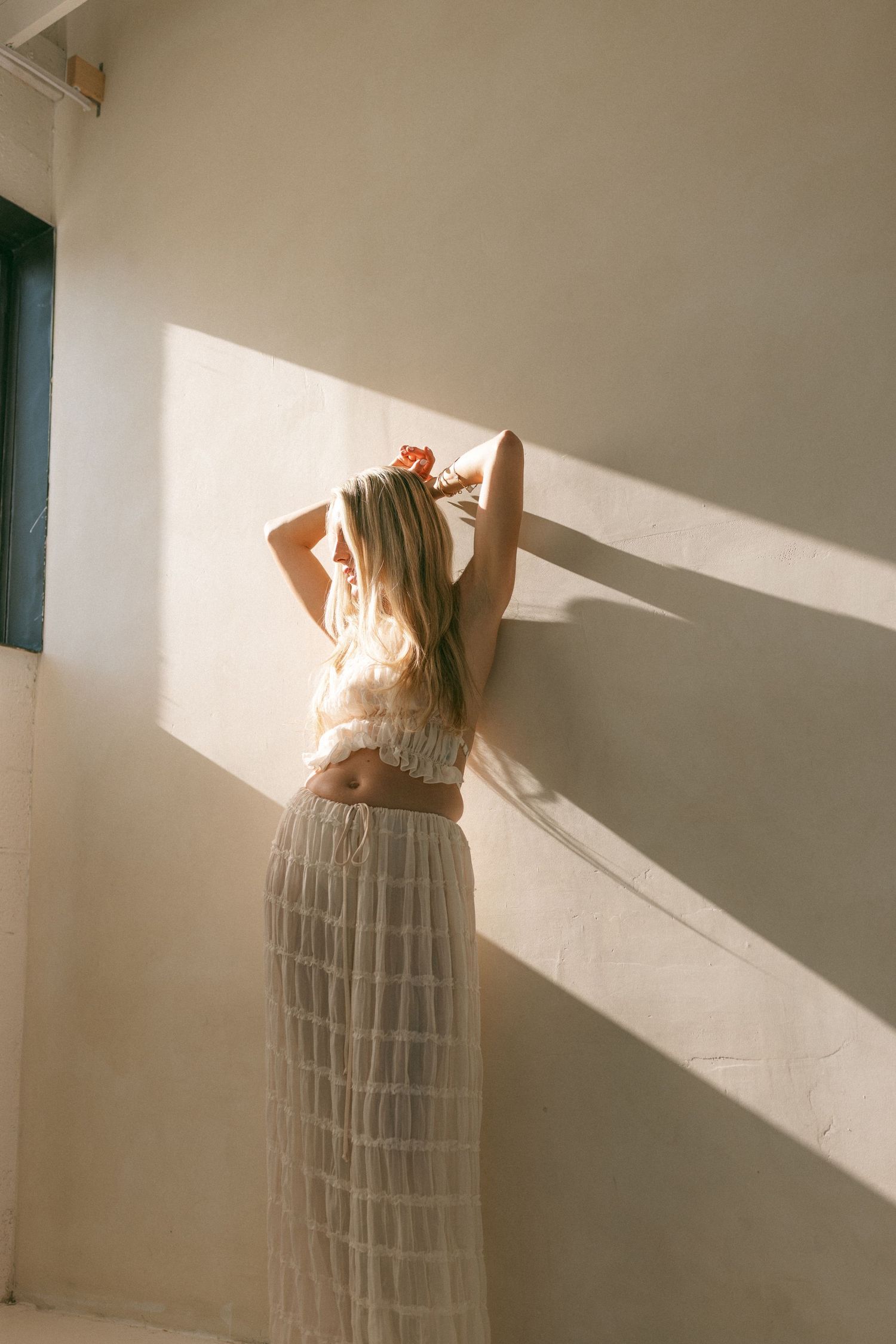 Blonde woman in sheer skirt and crop top poses against sunlit white wall with hands raised in her hair.
