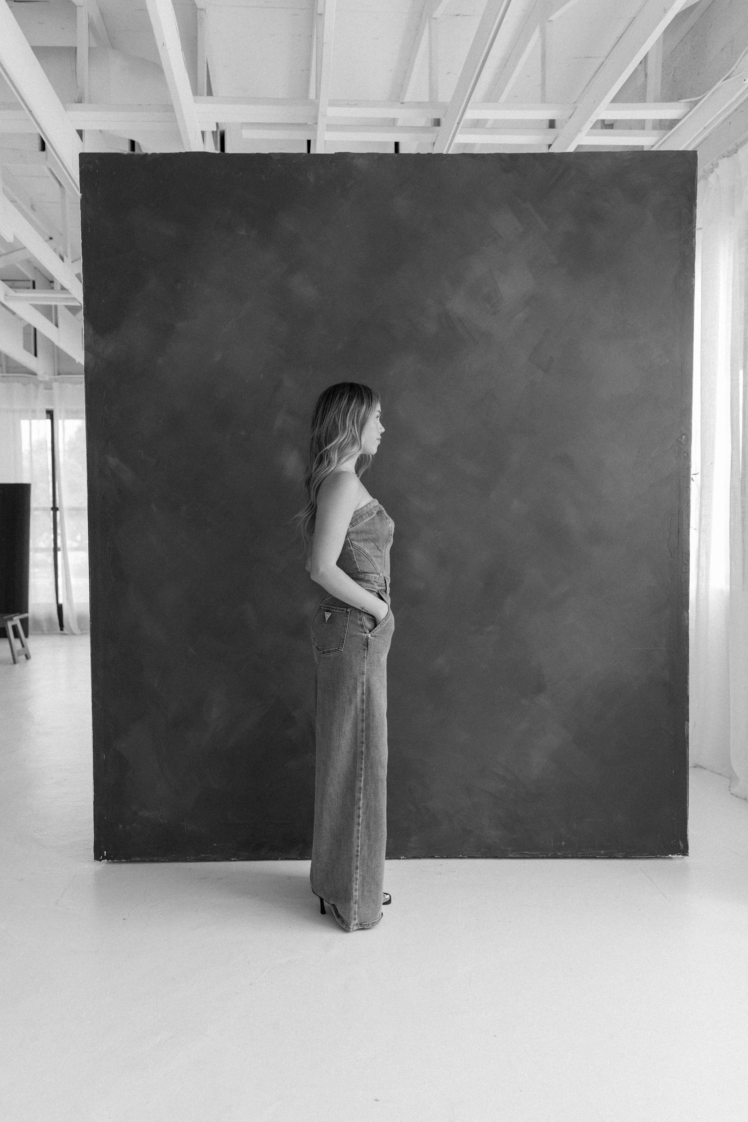 Black and white photo of a woman standing sideways in wide-leg jeans before a dark textured backdrop in a studio.