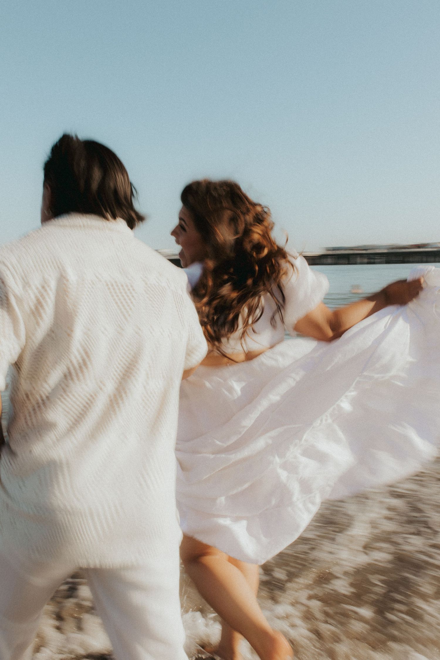 A couple in white attire runs joyfully along a sunny beach, her dress flowing in the breeze.