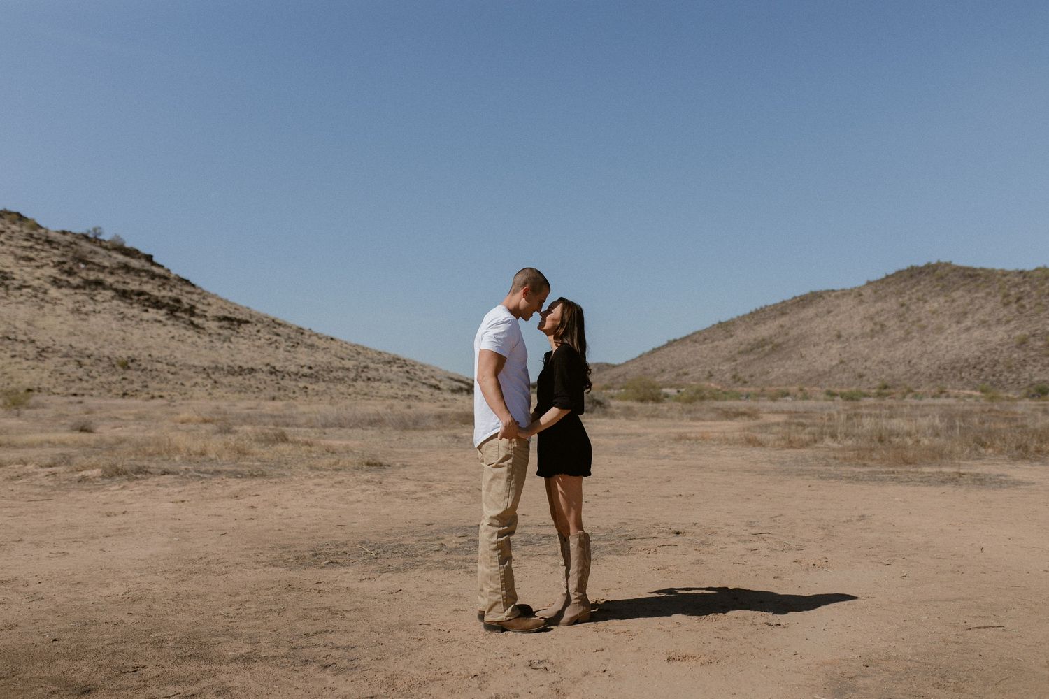 A couple shares an intimate moment in a desert landscape with rolling hills under bright blue sky.