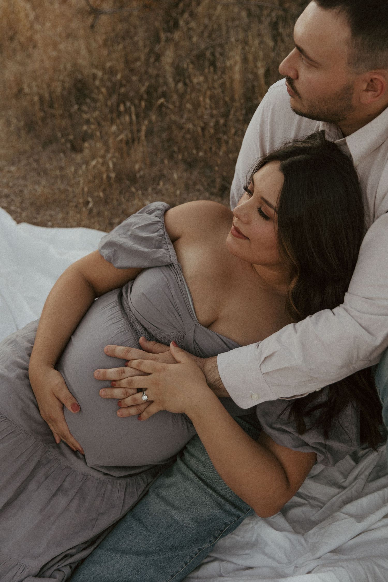 A tender maternity moment captured as a couple reclines on white blankets outdoors.