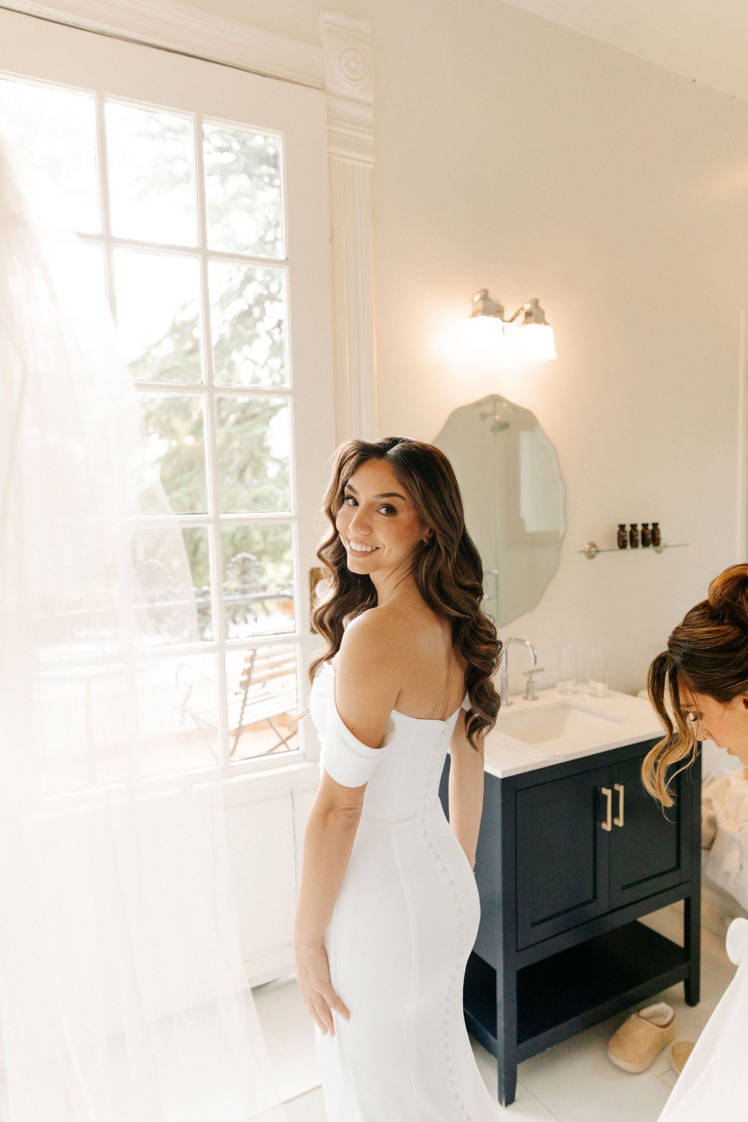 A bride in a white dress gets ready in a bright sunlit room with modern decor and navy blue furniture.