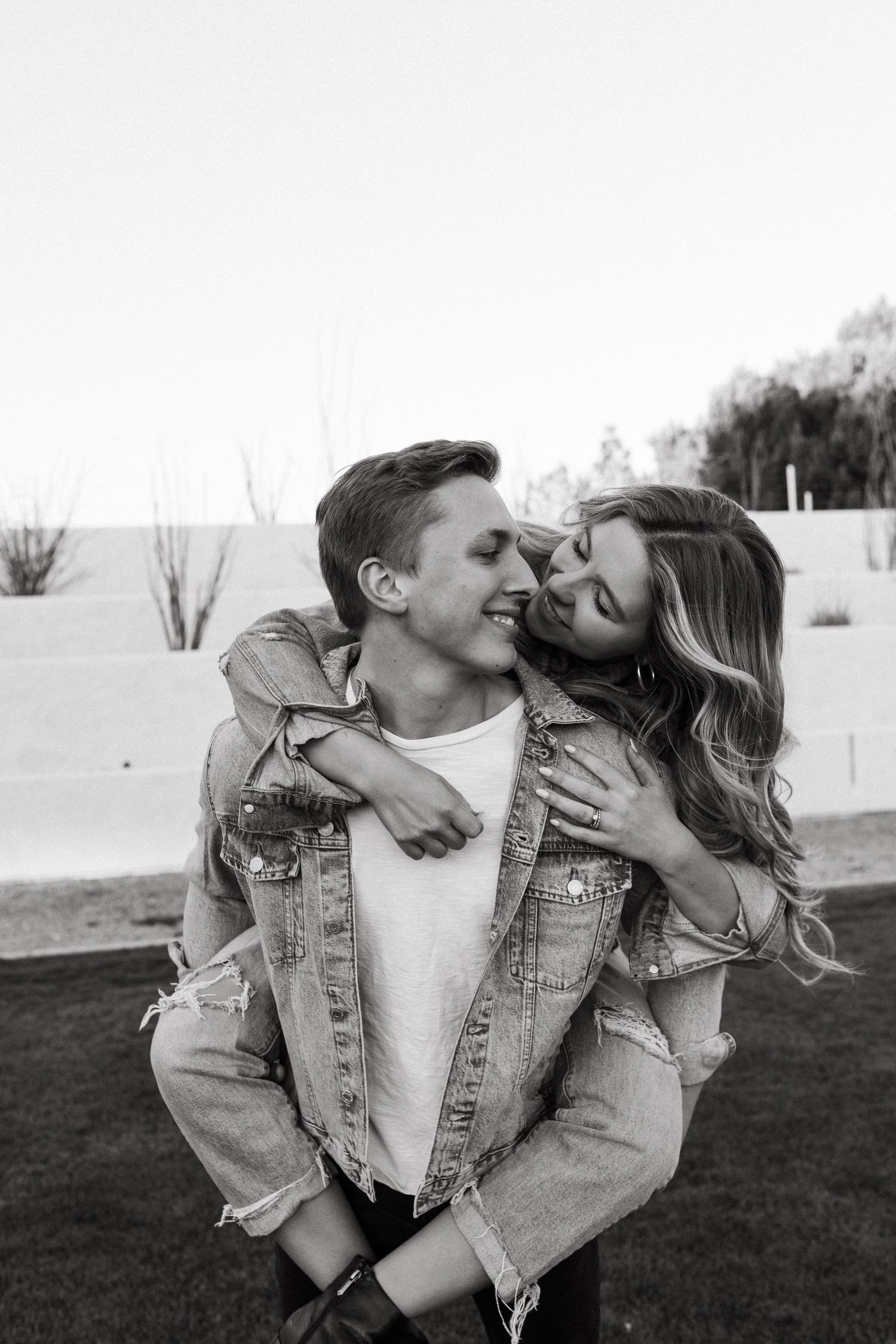 A black and white portrait of a couple in denim jackets sharing a playful piggyback moment.