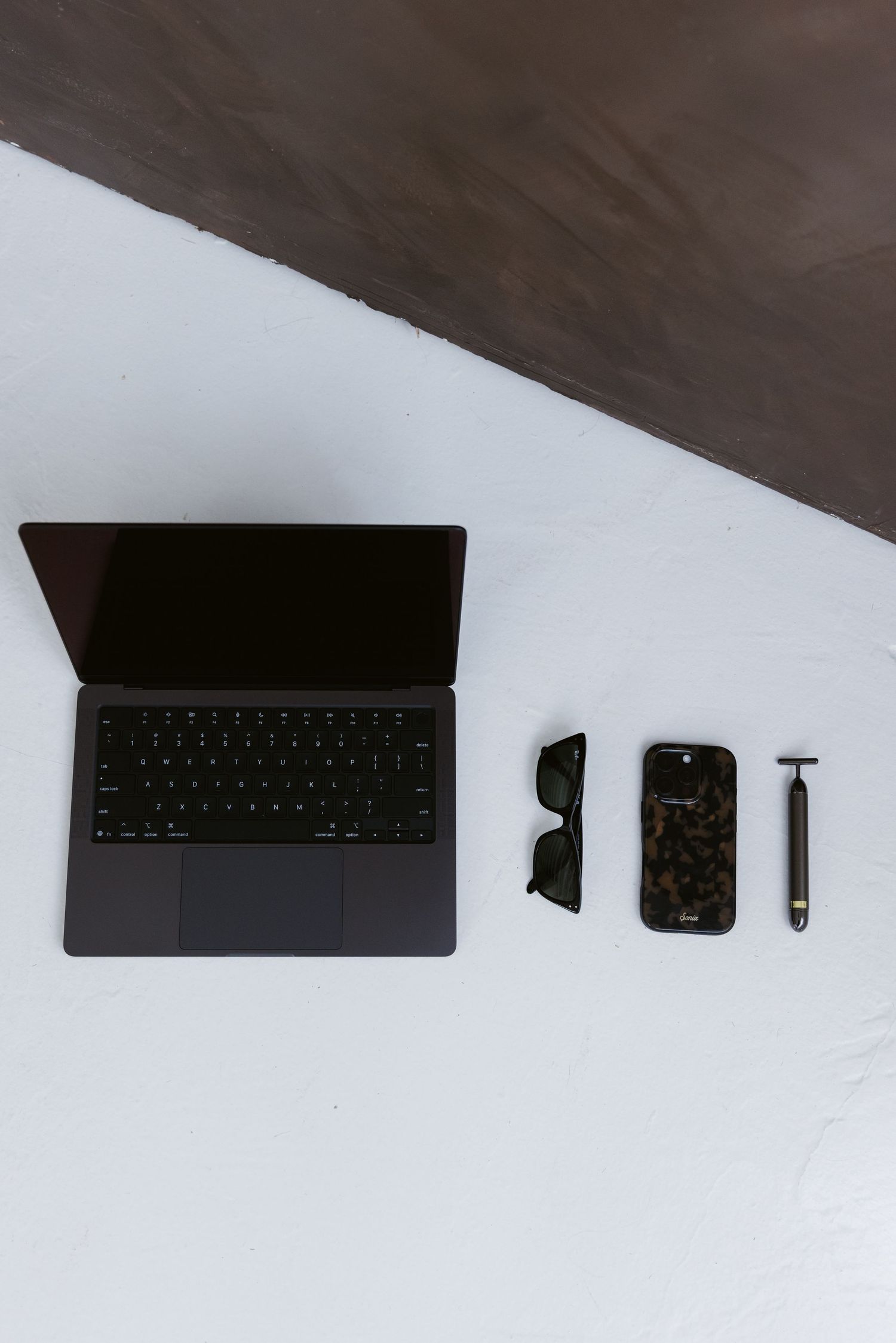 Flat lay of MacBook laptop, sunglasses, phone case, and razor on white surface.