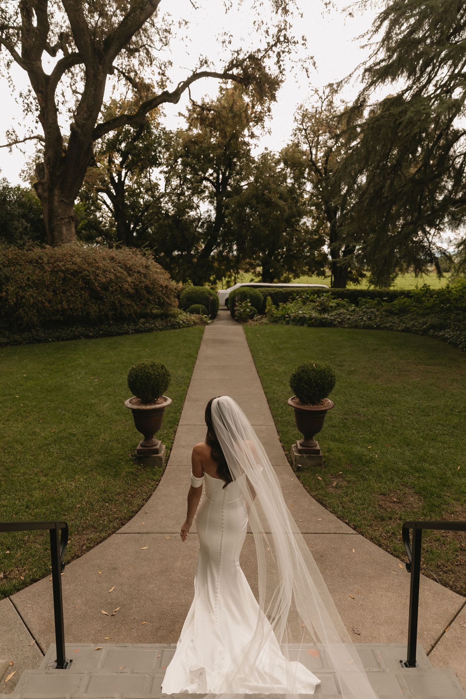 A long white veil flows behind someone walking down a tree-lined stone pathway in a garden setting.