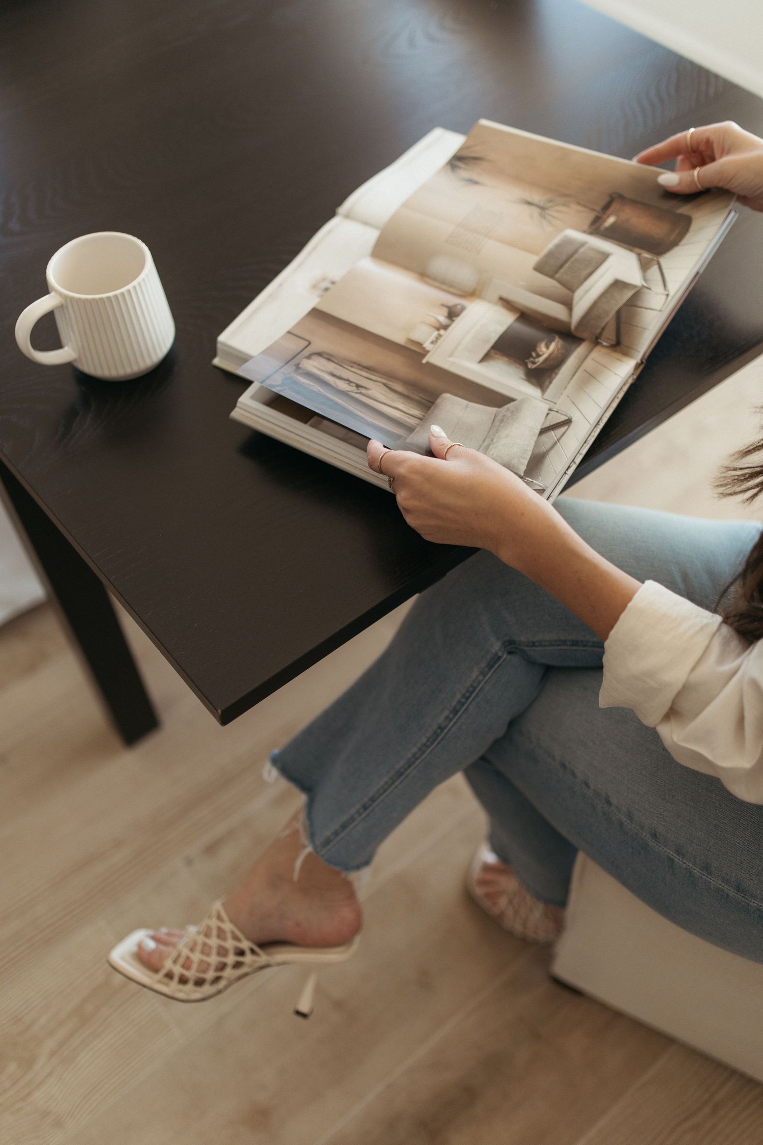 Person reading a magazine on a dark surface with a white coffee cup nearby while wearing casual attire.