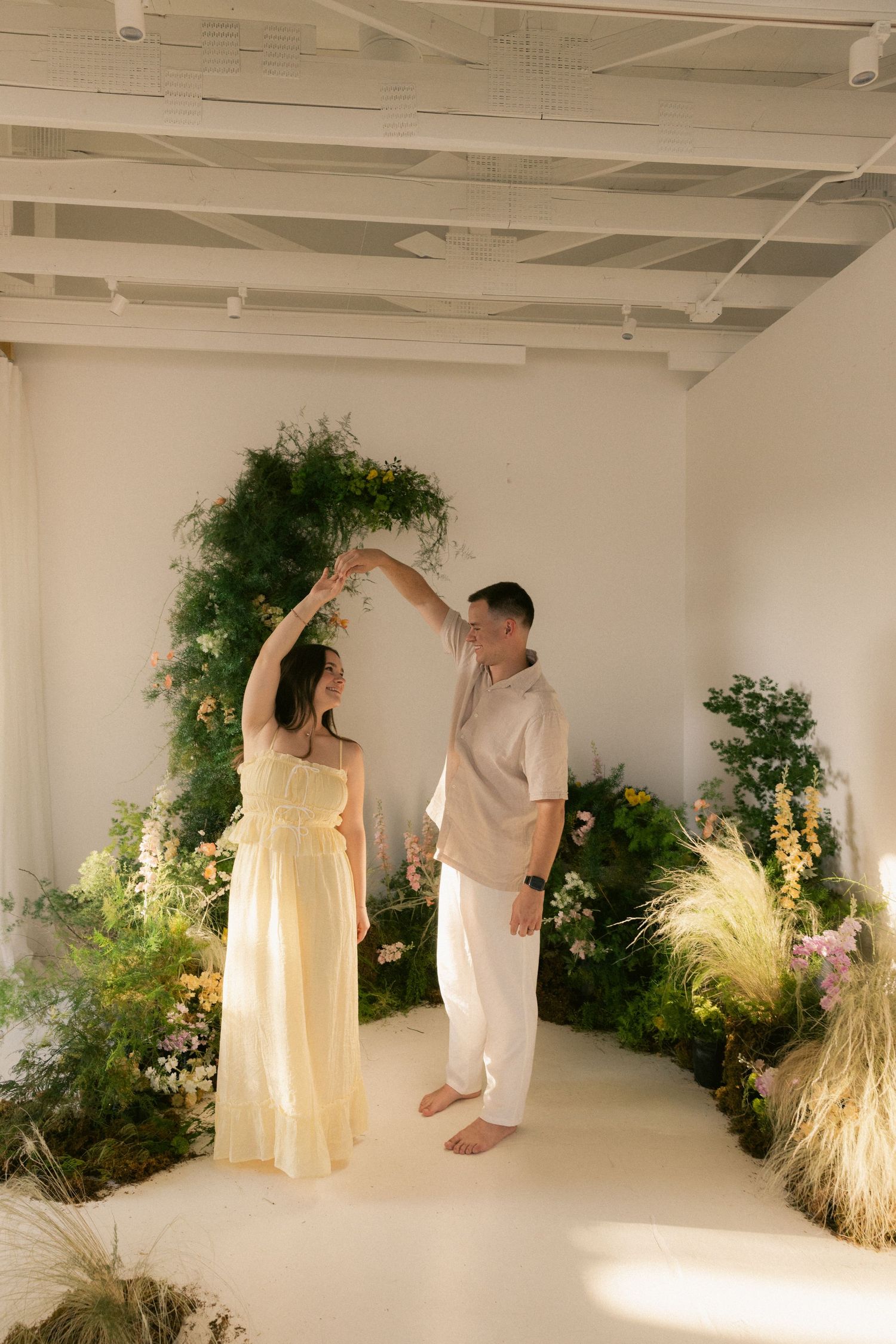 Couple dancing together in a bright white studio surrounded by lush green plants and colorful flowers.