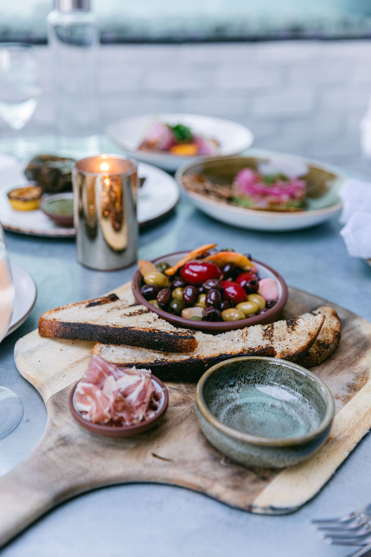 Rustic bread board with appetizers, olives, and small plates set on a blue tablecloth.