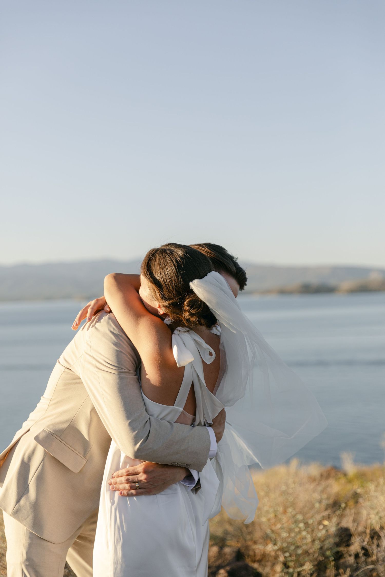 A romantic embrace between newlyweds against a waterfront backdrop at sunset.